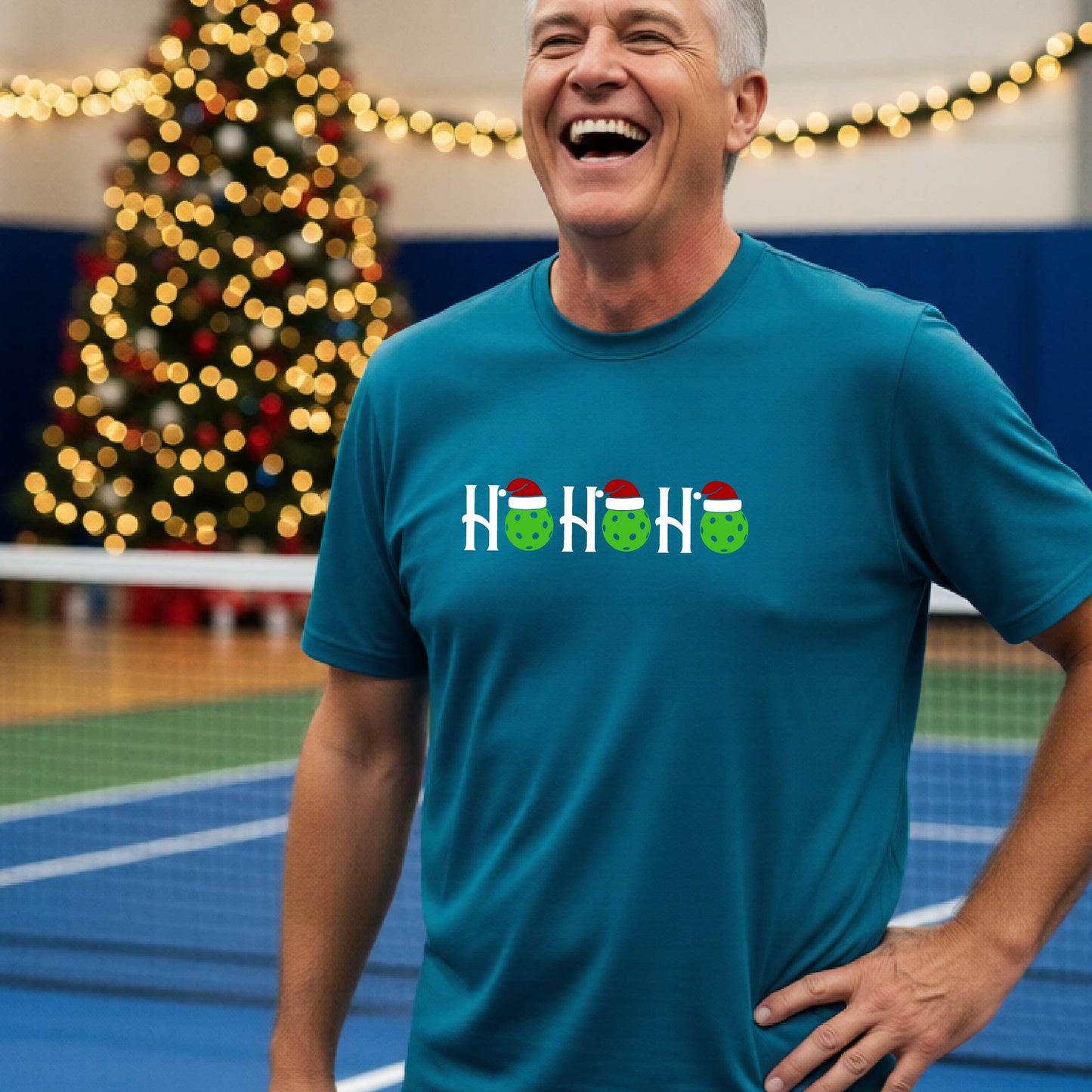 A man wearing a blue short-sleeve t-shirt with a 'HO HO HO' graphic made of Christmas pickleballs wearing red Santa hats, set against a blurred background of a pickleball court and a decorated Christmas tree.