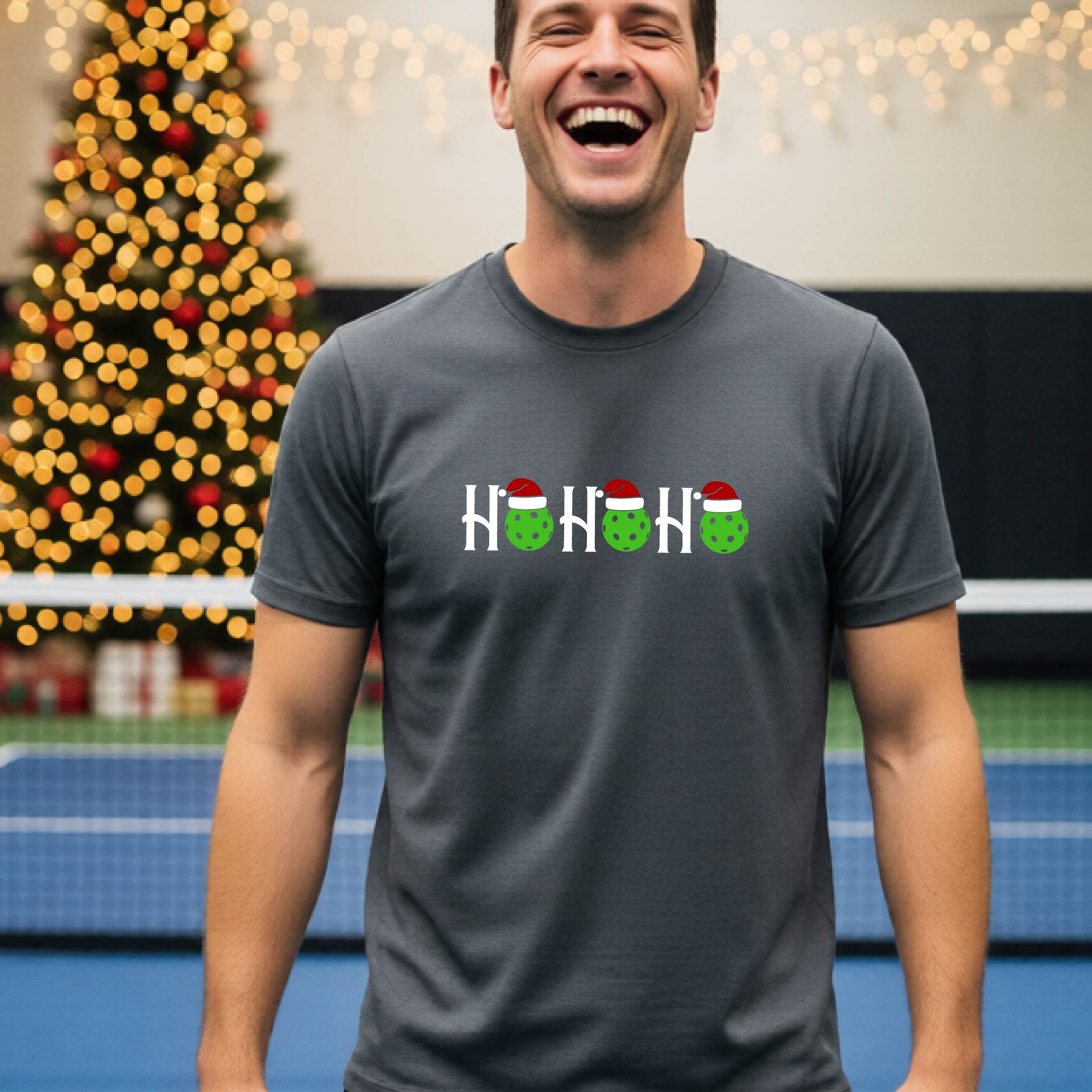 A man wearing a gray short-sleeve t-shirt with a 'HO HO HO' graphic made of Christmas pickleballs wearing red Santa hats, set against a blurred background of a pickleball court and a decorated Christmas tree.