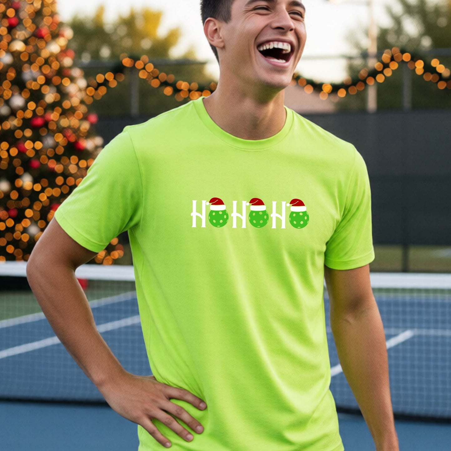 A man wearing a lime green short-sleeve t-shirt with a 'HO HO HO' graphic made of Christmas pickleballs wearing red Santa hats, set against a blurred background of a pickleball court and a decorated Christmas tree.