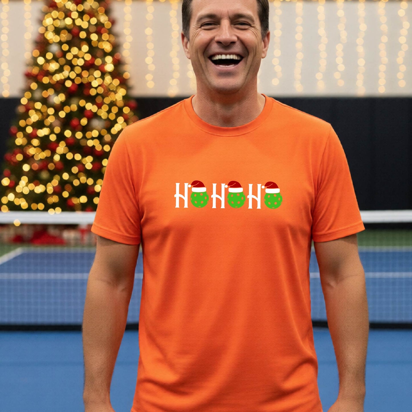 A man wearing a bright orange short-sleeve t-shirt with a 'HO HO HO' graphic made of Christmas pickleballs wearing red Santa hats, set against a blurred background of a pickleball court and a decorated Christmas tree.