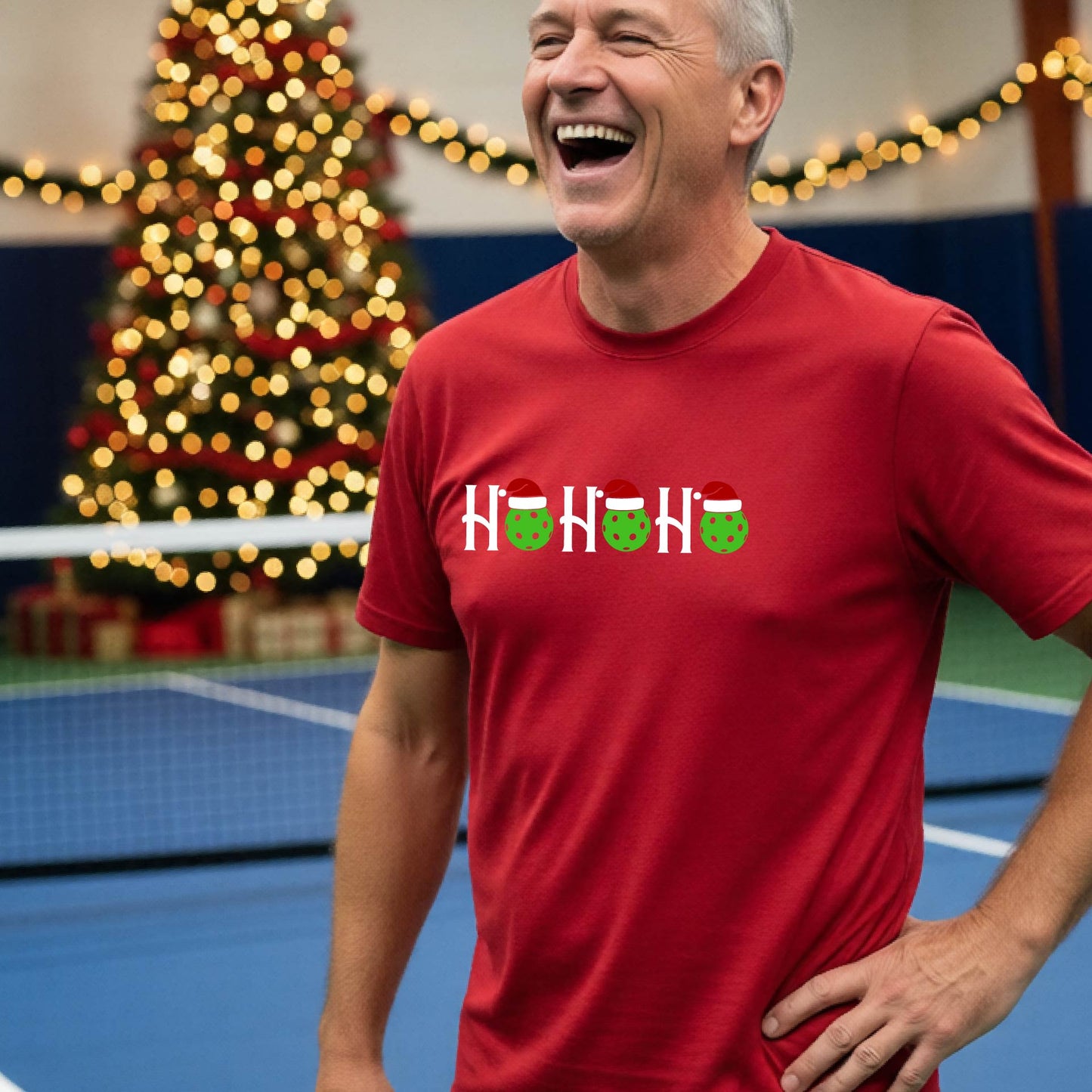 A man wearing a red short-sleeve t-shirt with a 'HO HO HO' graphic made of Christmas pickleballs wearing red Santa hats, set against a blurred background of a pickleball court and a decorated Christmas tree.