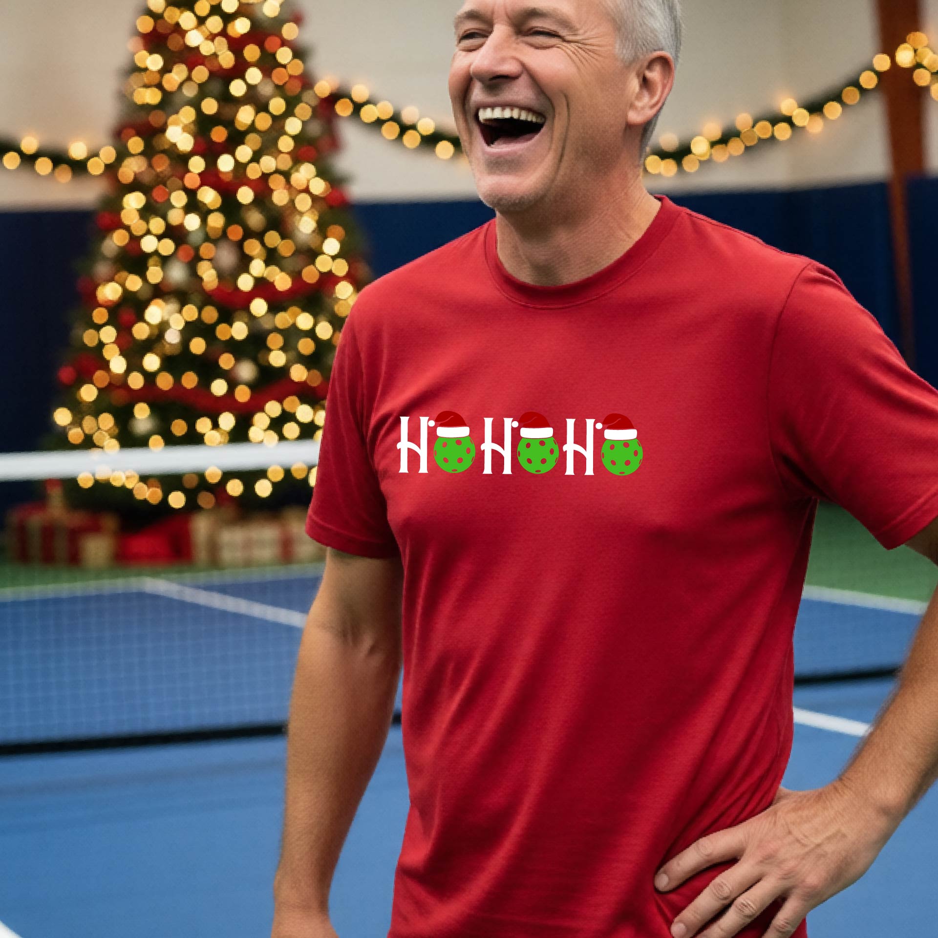 A man wearing a red short-sleeve t-shirt with a 'HO HO HO' graphic made of Christmas pickleballs wearing red Santa hats, set against a blurred background of a pickleball court and a decorated Christmas tree.