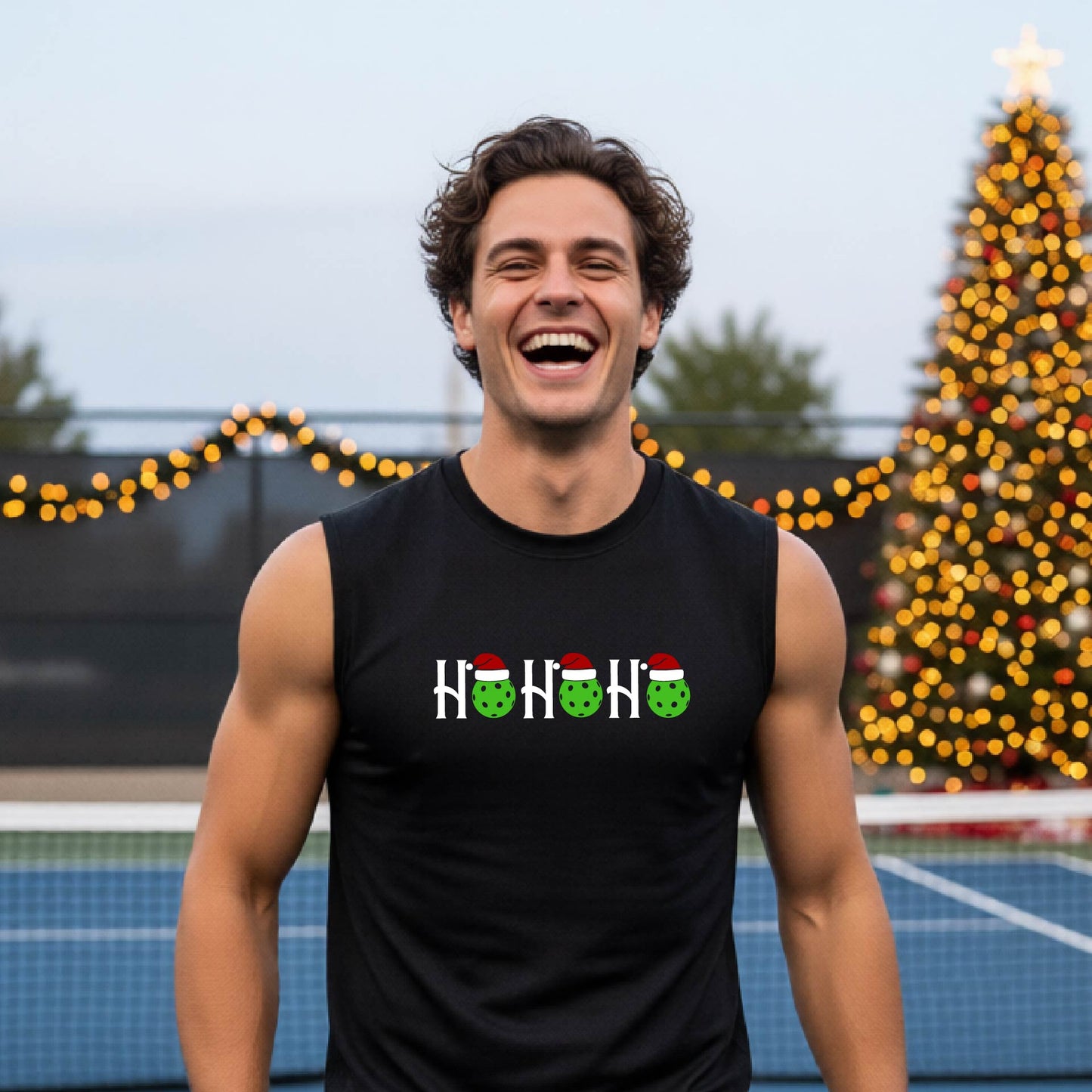 A man laughing on an  pickleball court wearing a black sleeveless shirt with a 'HO HO HO' holiday graphic made of green pickleballs wearing Santa hats. A decorated Christmas tree and warm string lights are blurred in the background.