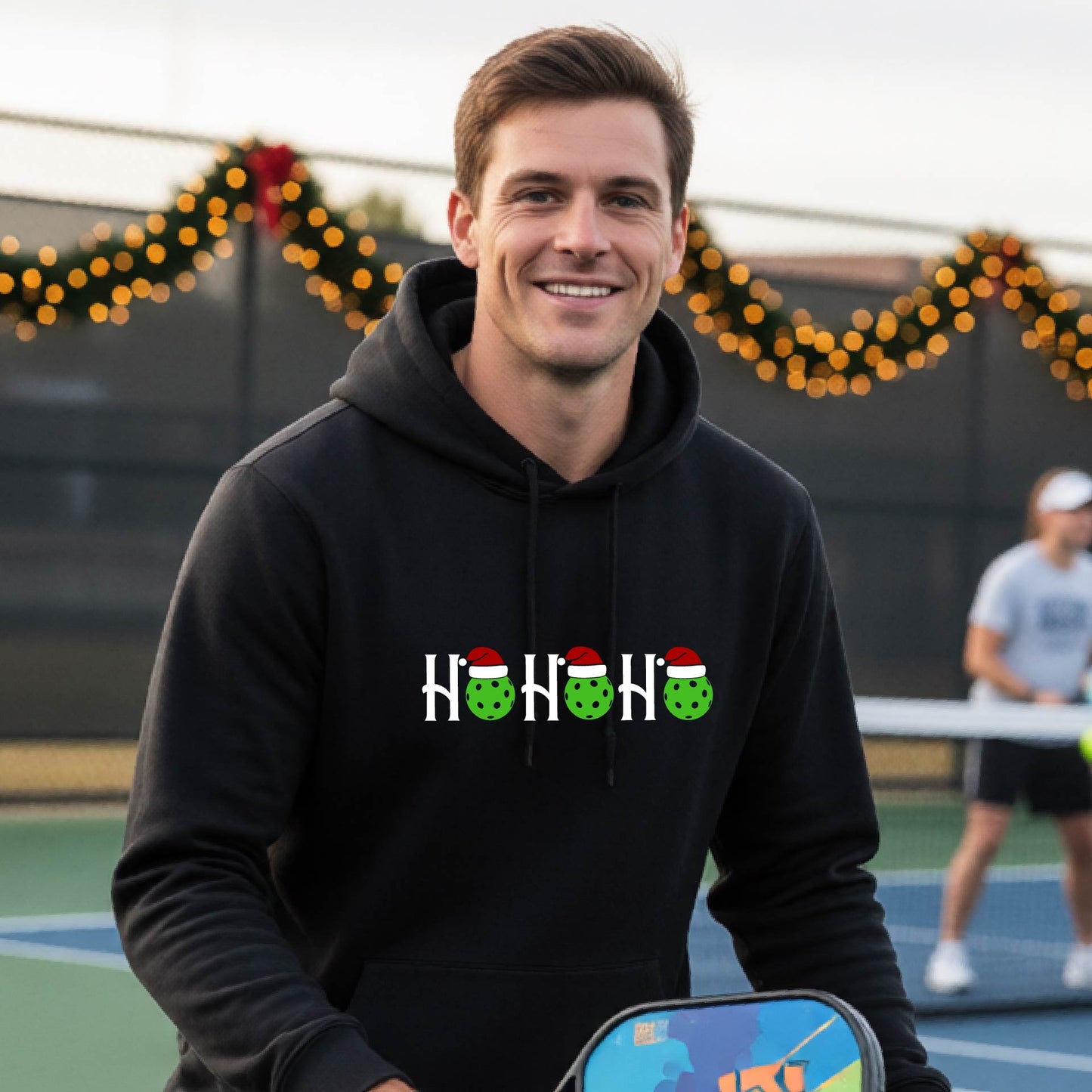 A man wearing a black hoodie with a 'HOHOHO' graphic made of green pickleballs  wearing Santa hats, set against a blurred  pickleball court background with holiday lights.
