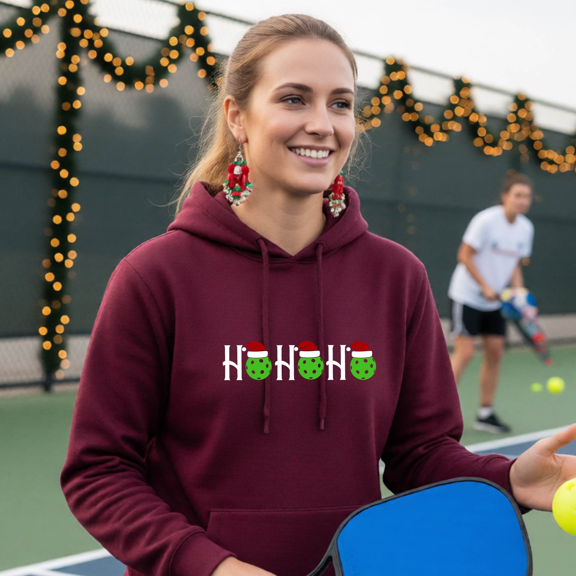 A woman wearing a maroon hoodie with a 'HOHOHO' graphic made of green pickleballs  wearing Santa hats, set against a blurred  pickleball court background with holiday lights.