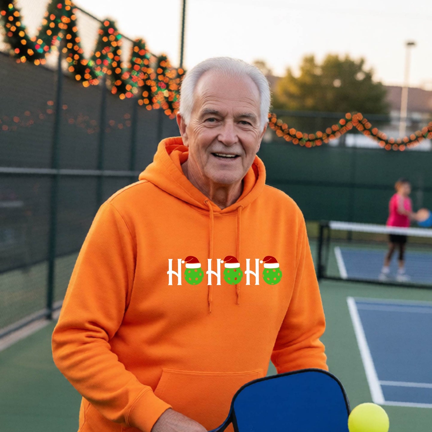 A man wearing a bright orange hoodie with a 'HOHOHO' graphic made of green pickleballs  wearing Santa hats, set against a blurred  pickleball court background with holiday lights.