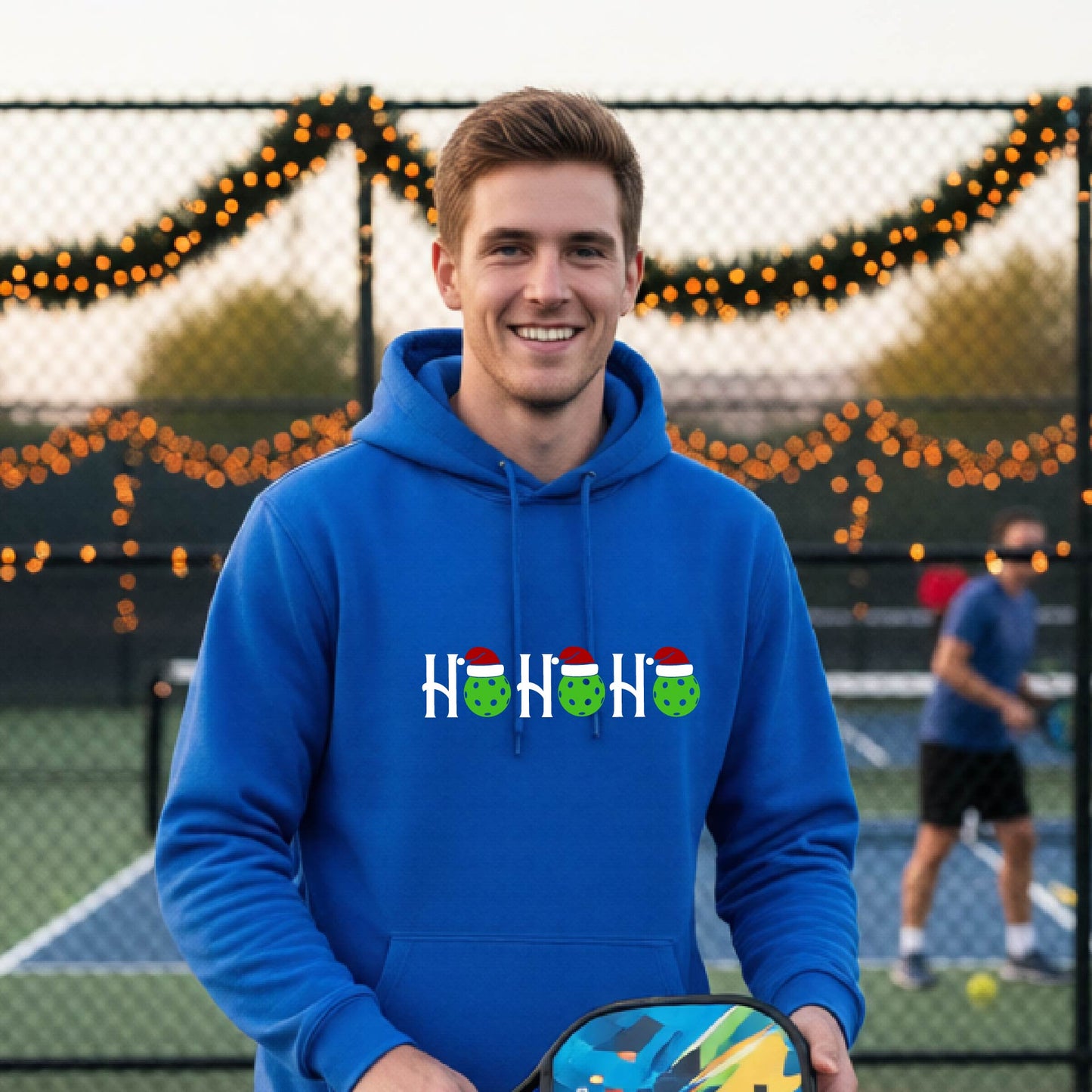 A man wearing a royal blue hoodie with a 'HOHOHO' graphic made of green pickleballs  wearing Santa hats, set against a blurred  pickleball court background with holiday lights.