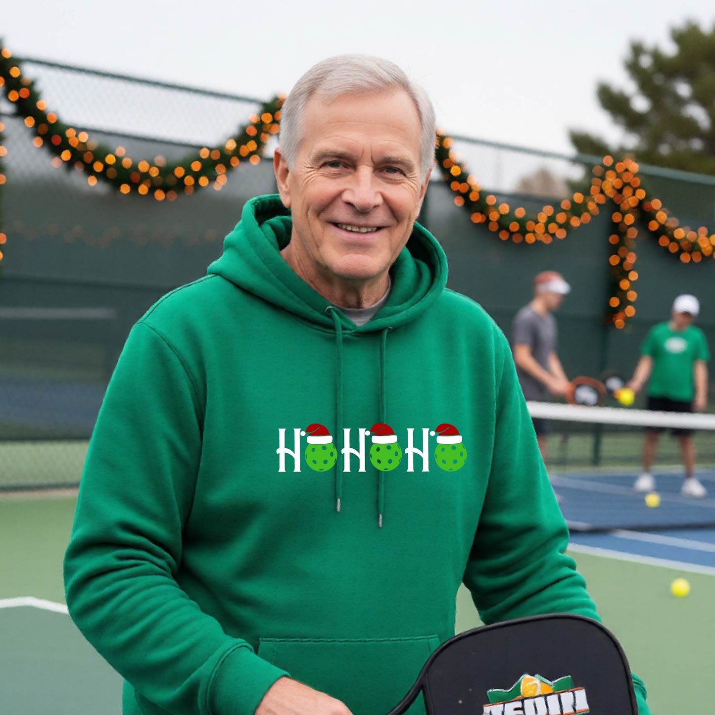 A man wearing a green hoodie with a 'HOHOHO' graphic made of green pickleballs  wearing Santa hats, set against a blurred  pickleball court background with holiday lights.