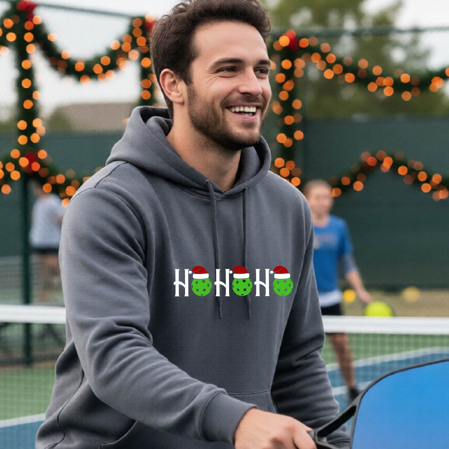 A man wearing a dark gray hoodie with a 'HOHOHO' graphic made of green pickleballs  wearing Santa hats, set against a blurred  pickleball court background with holiday lights.