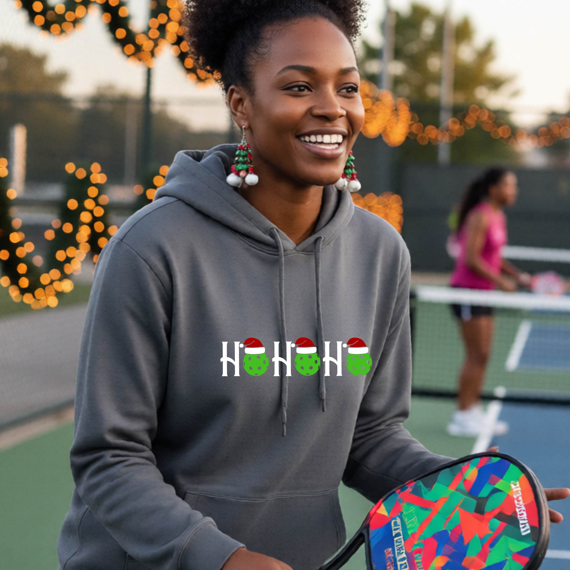 A womman wearing a dark gray hoodie with a 'HOHOHO' graphic made of green pickleballs  wearing Santa hats, set against a blurred  pickleball court background with holiday lights.