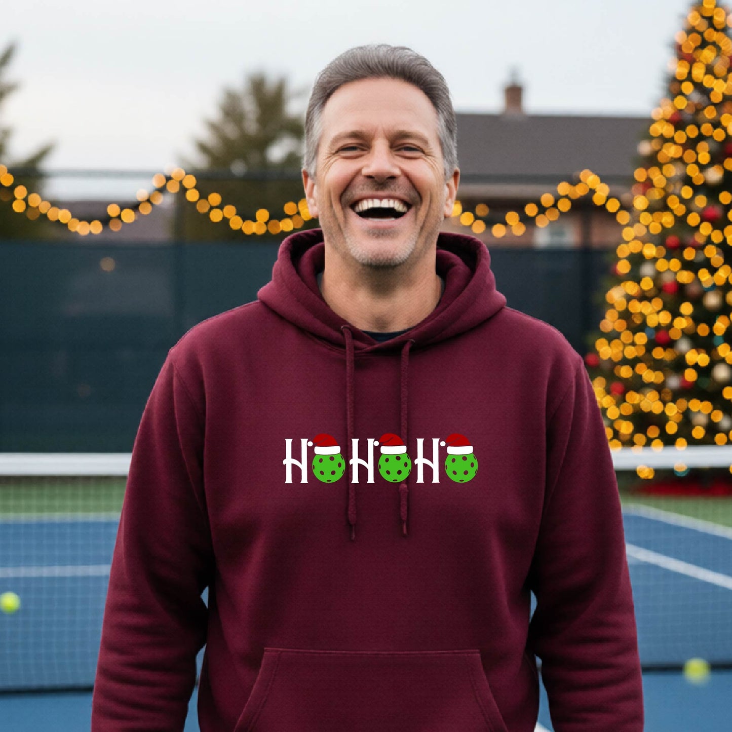 A man wearing a maroon hoodie with a 'HOHOHO' graphic made of green pickleballs  wearing Santa hats, set against a blurred  pickleball court background with holiday lights.