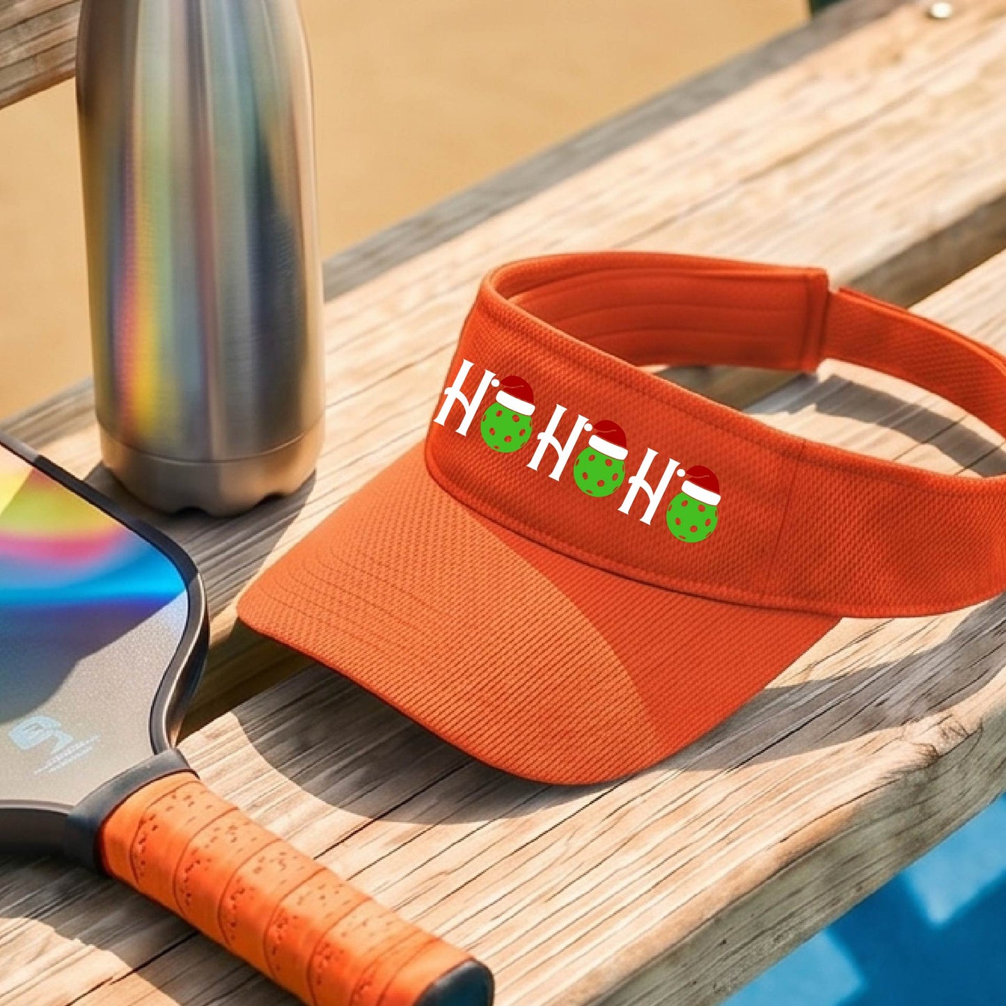 An orange visor with a pickleball Christmas graphic of the text "HO HO HO" with the O replaced by green picklebalsl paddle  santa hats. 