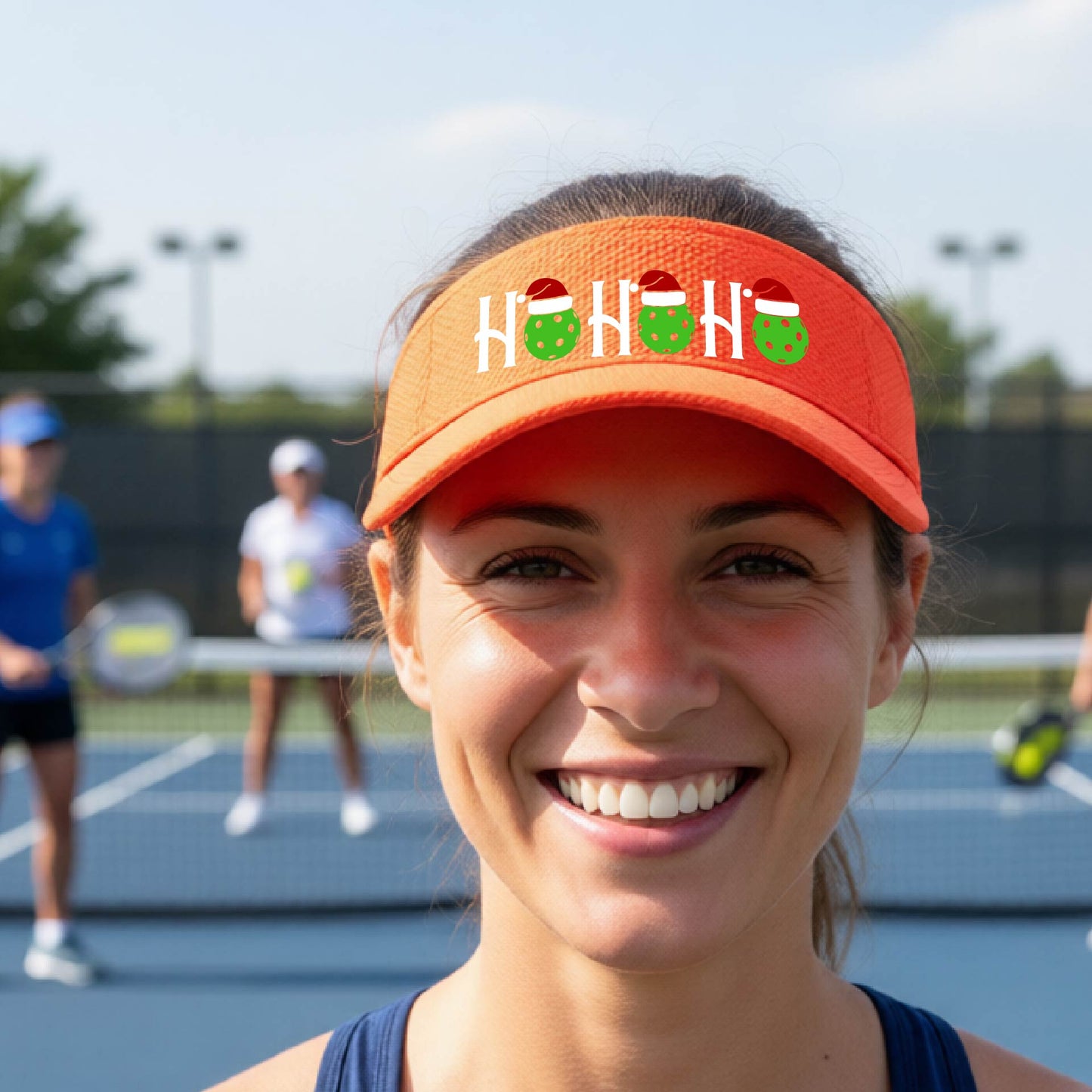 An orange visor with a pickleball Christmas graphic of the text "HO HO HO" with the O replaced by green picklebalsl paddle  santa hats. 