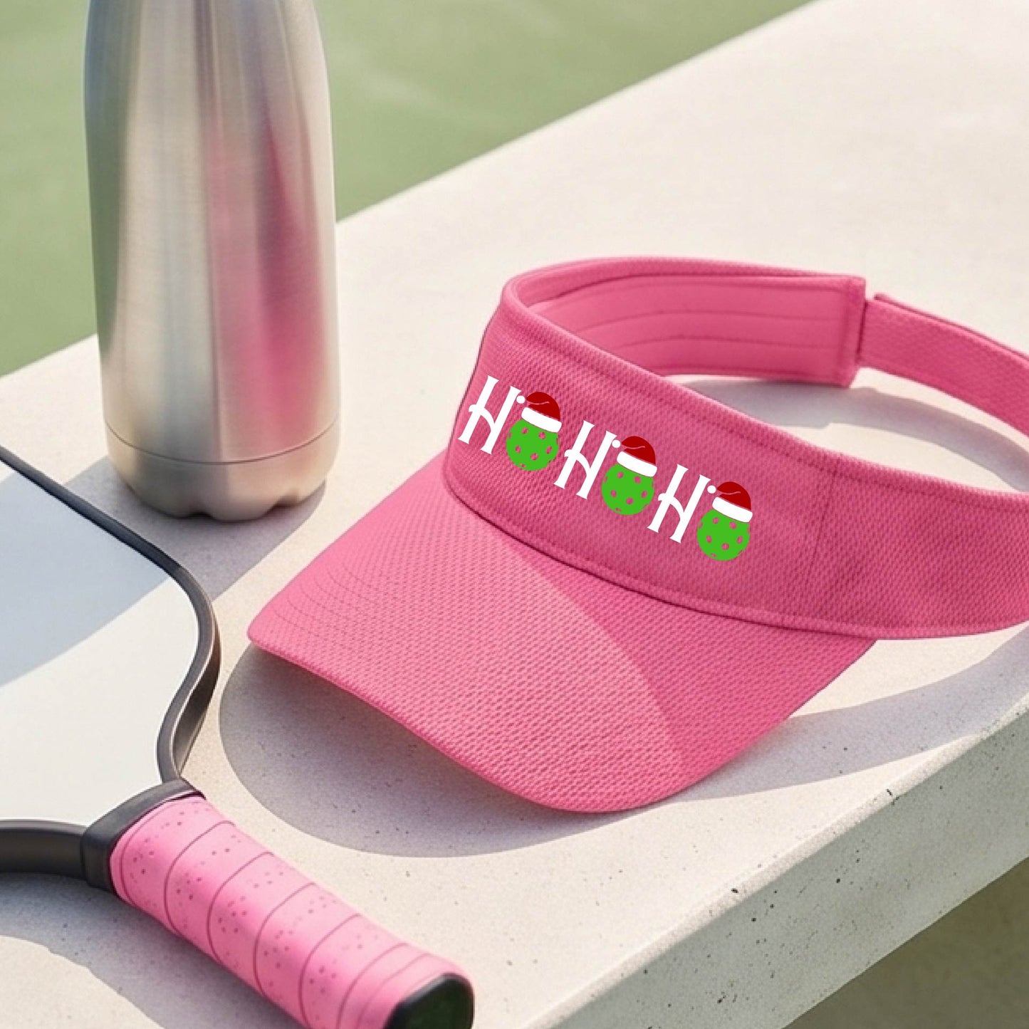 A pink visor with a pickleball Christmas graphic of the text "HO HO HO" with the O replaced by green picklebalsl paddle  santa hats. 