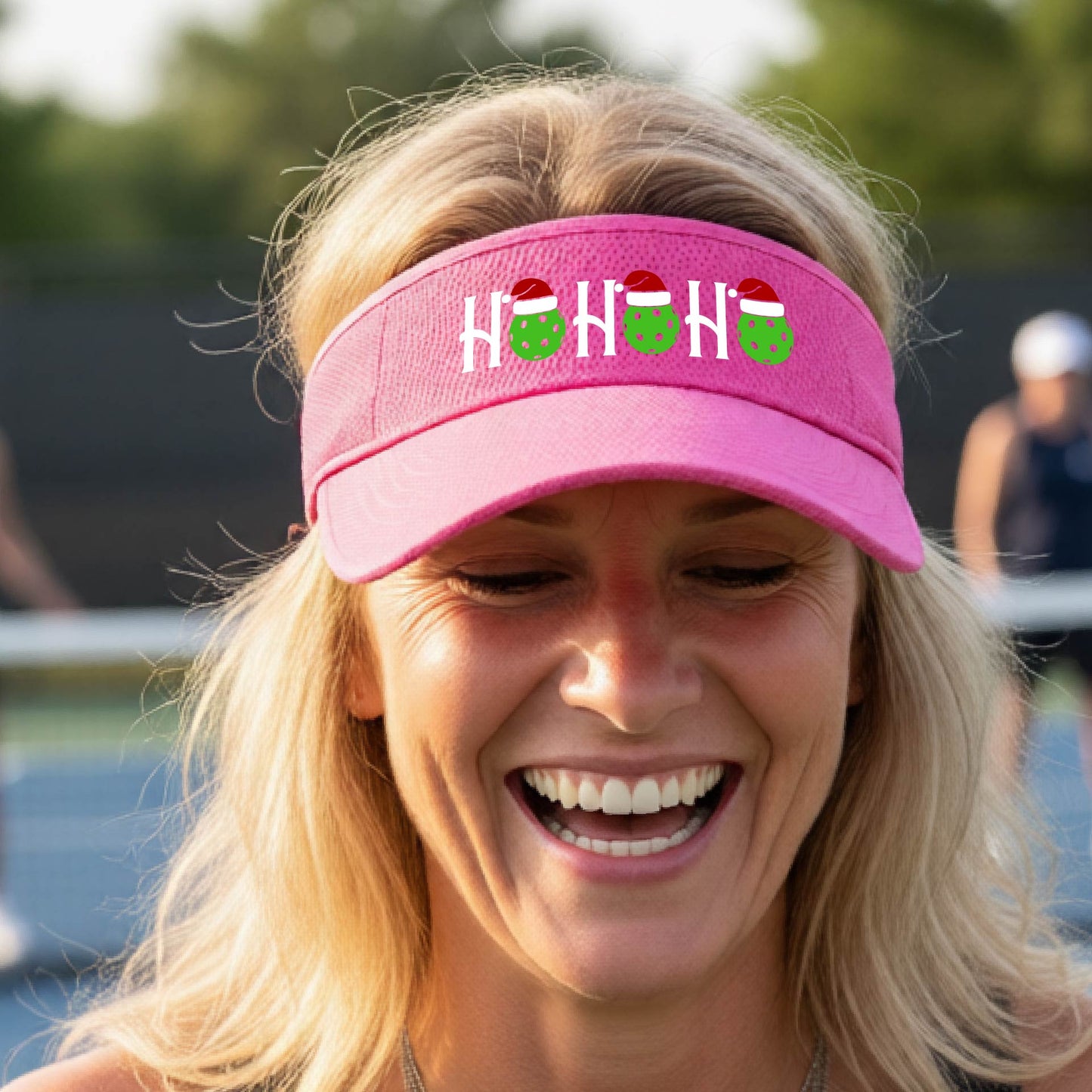 A pink visor with a pickleball Christmas graphic of the text "HO HO HO" with the O replaced by green picklebalsl paddle  santa hats. 