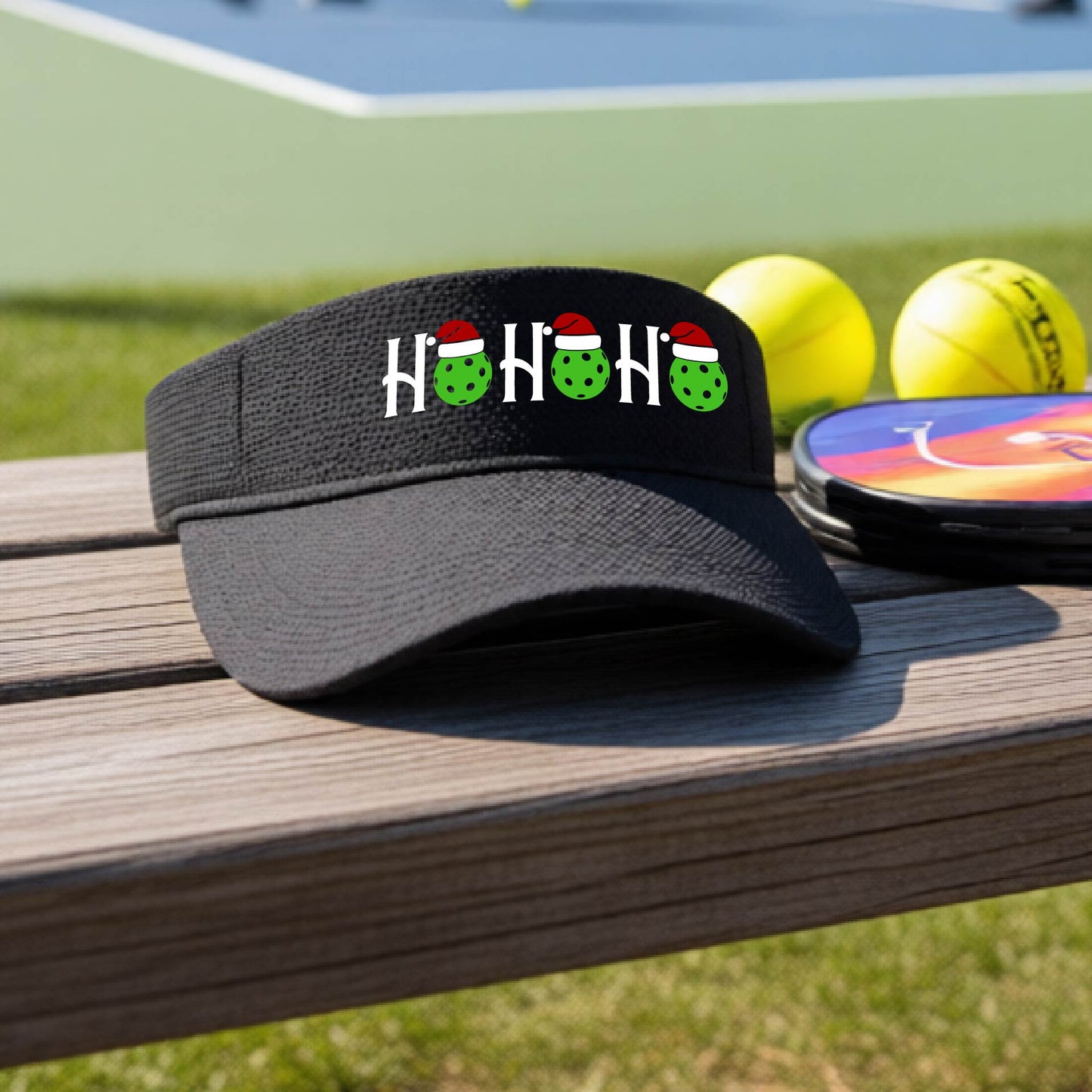 A black visor with a pickleball Christmas graphic of the text "HO HO HO" with the O replaced by green picklebalsl paddle  santa hats. 