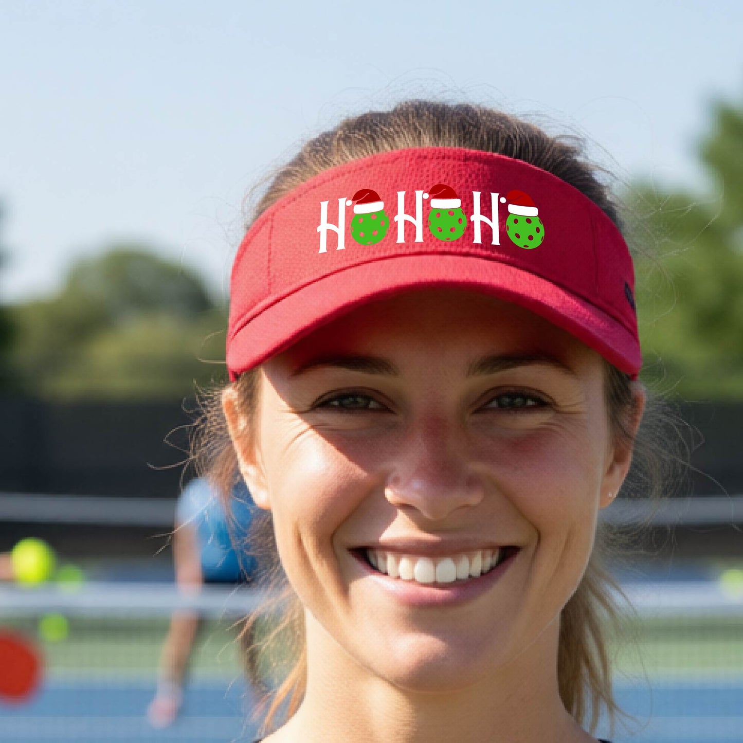 A red visor with a pickleball Christmas graphic of the text "HO HO HO" with the O replaced by green picklebalsl paddle  santa hats. 