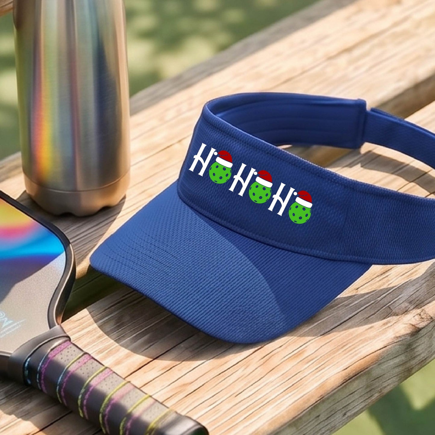 A royal blue visor with a pickleball Christmas graphic of the text "HO HO HO" with the O replaced by green picklebalsl paddle  santa hats. 