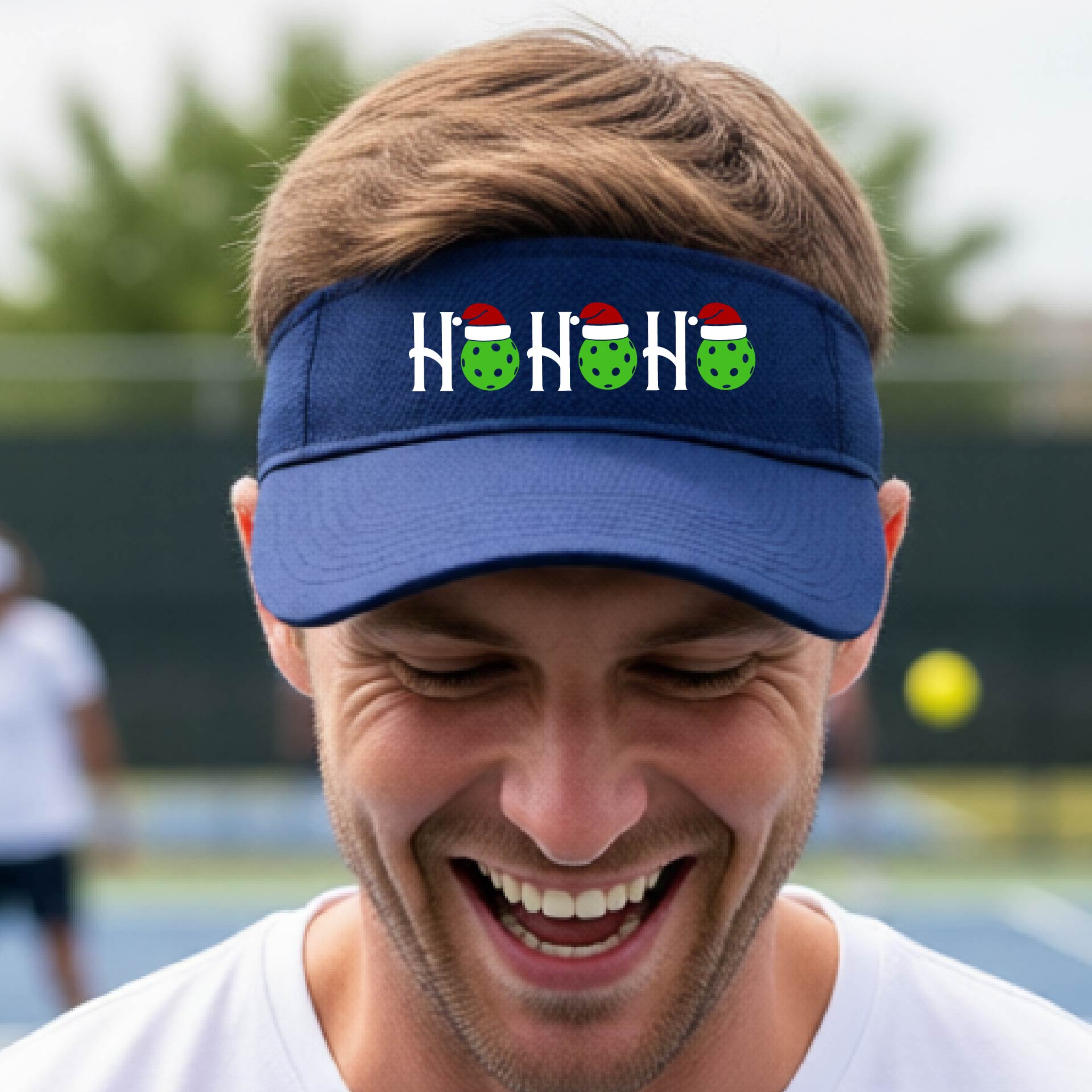 A royal blue visor with a pickleball Christmas graphic of the text "HO HO HO" with the O replaced by green picklebalsl paddle  santa hats. 
