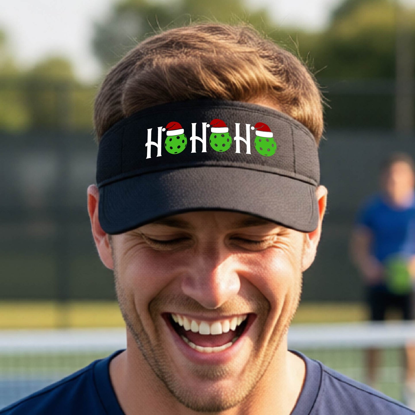 A black visor with a pickleball Christmas graphic of the text "HO HO HO" with the O replaced by green picklebalsl paddle  santa hats. 