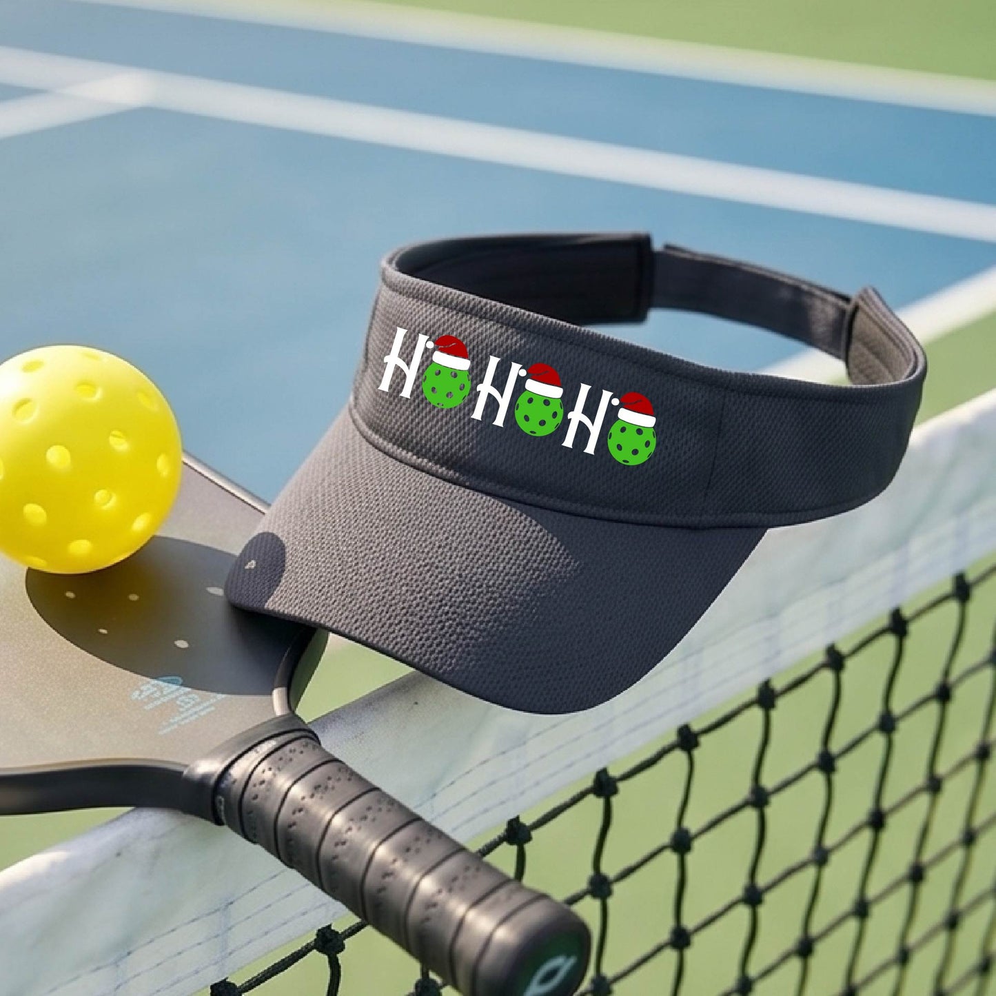 A graphite gray visor with a pickleball Christmas graphic of the text "HO HO HO" with the O replaced by green picklebalsl paddle  santa hats. 