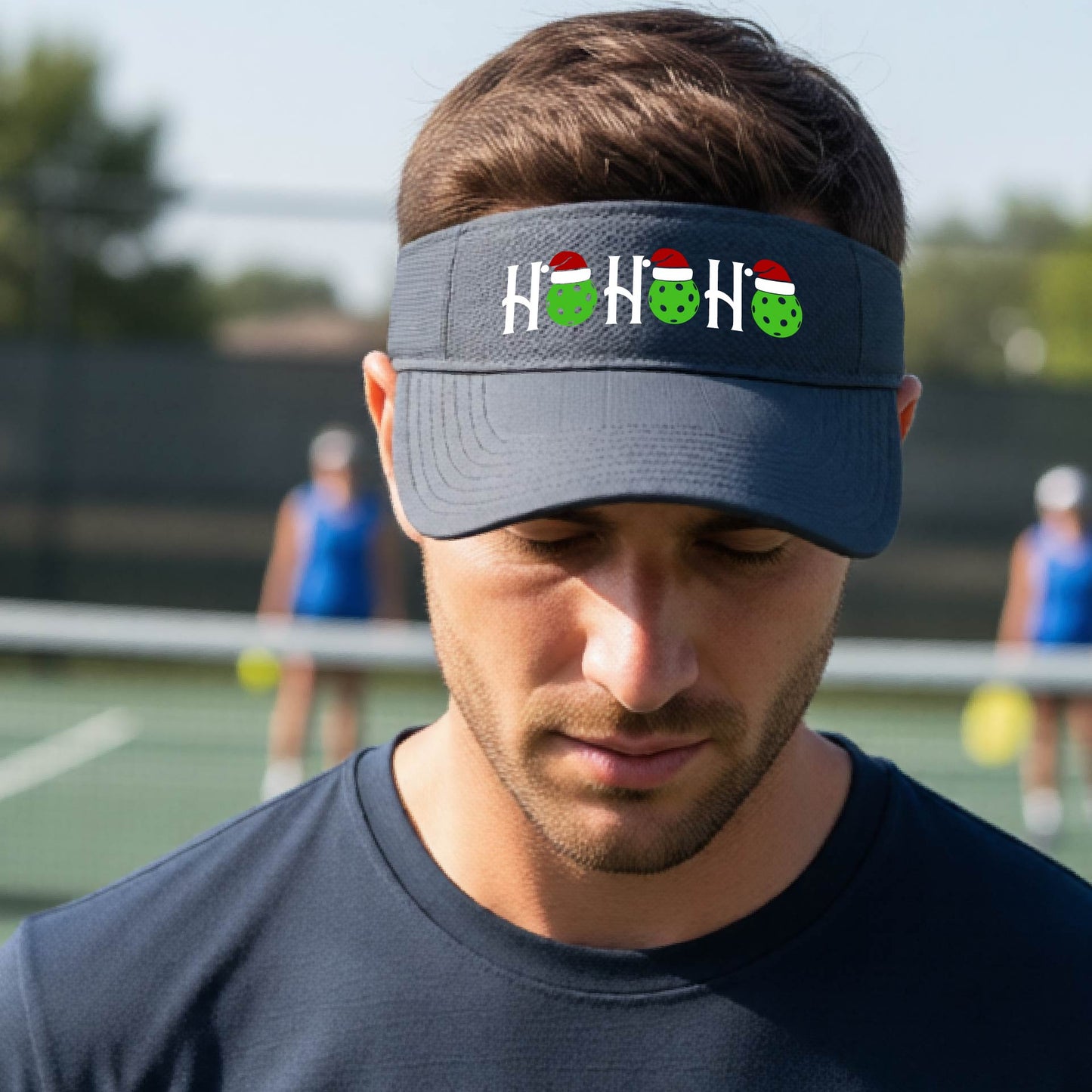 A graphite gray visor with a pickleball Christmas graphic of the text "HO HO HO" with the O replaced by green picklebalsl paddle  santa hats. 