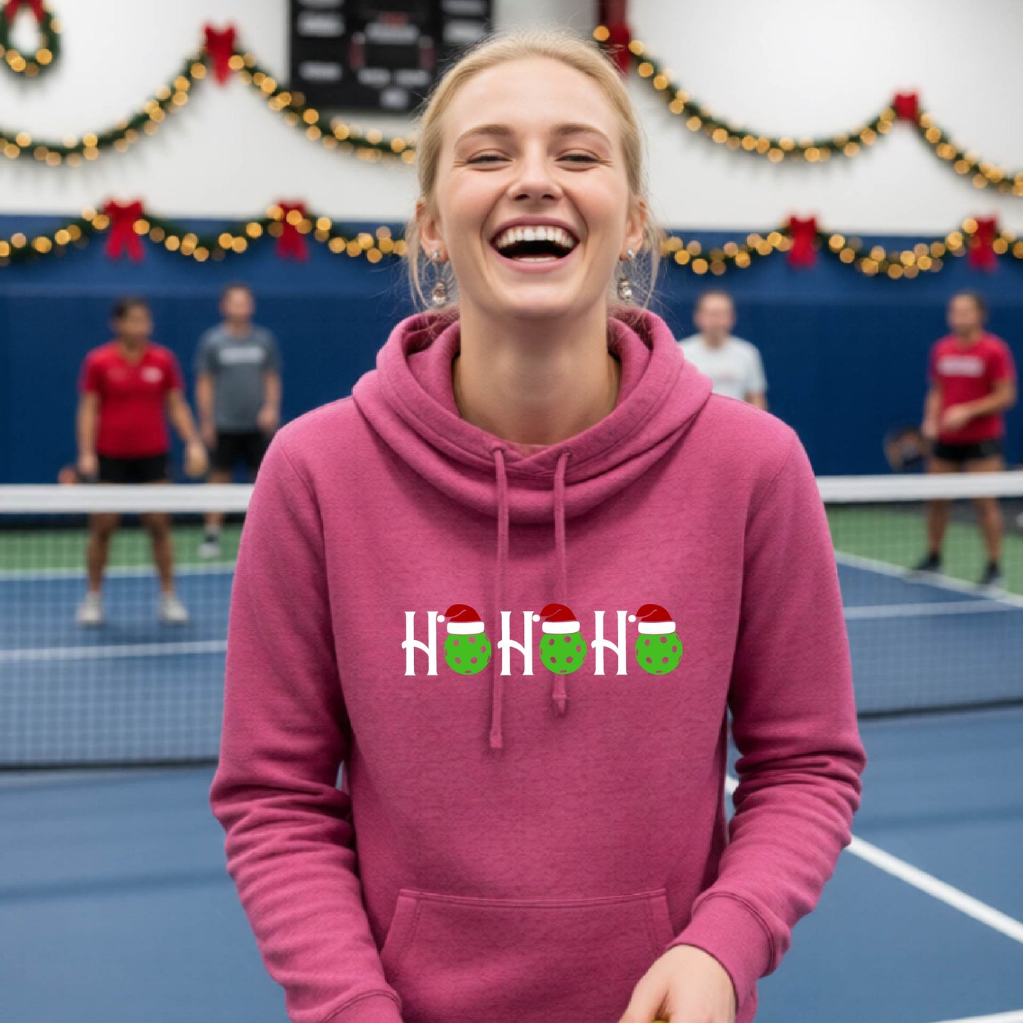 A pink cowl-neck hooded sweatshirt featuring a graphic print that reads "Ho Ho Ho'" in white text. The design includes pickleball replacing the O in the text.