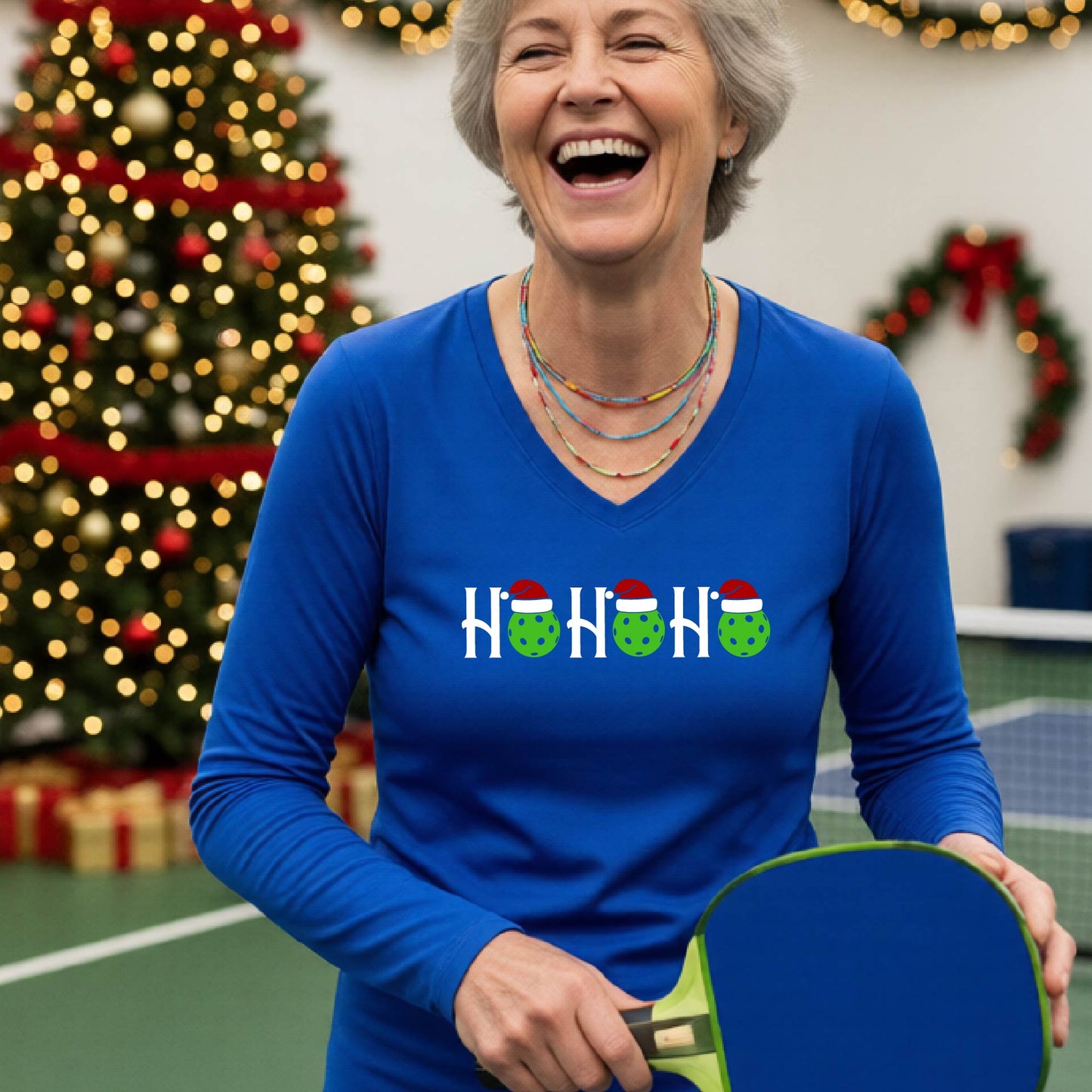 A women's royal blue long-sleeve v-neck shirt featuring a pickleball graphic with text "HO HO HO"  with the O replaced by green pickleballs wearing a santa hats and  Christmas themed background.