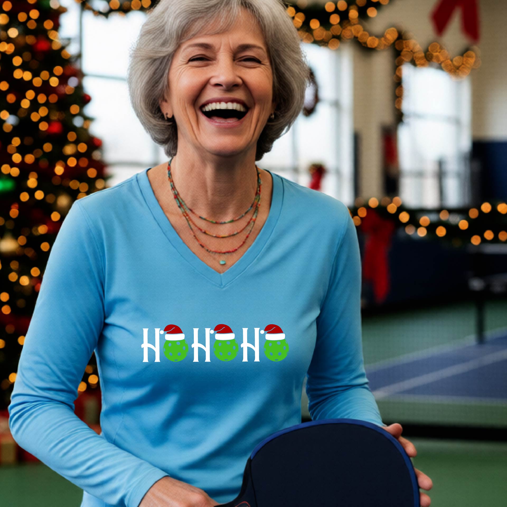 A women's light blue long-sleeve v-neck shirt featuring a pickleball graphic with text "HO HO HO"  with the O replaced by green pickleballs wearing a santa hats and  Christmas themed background.