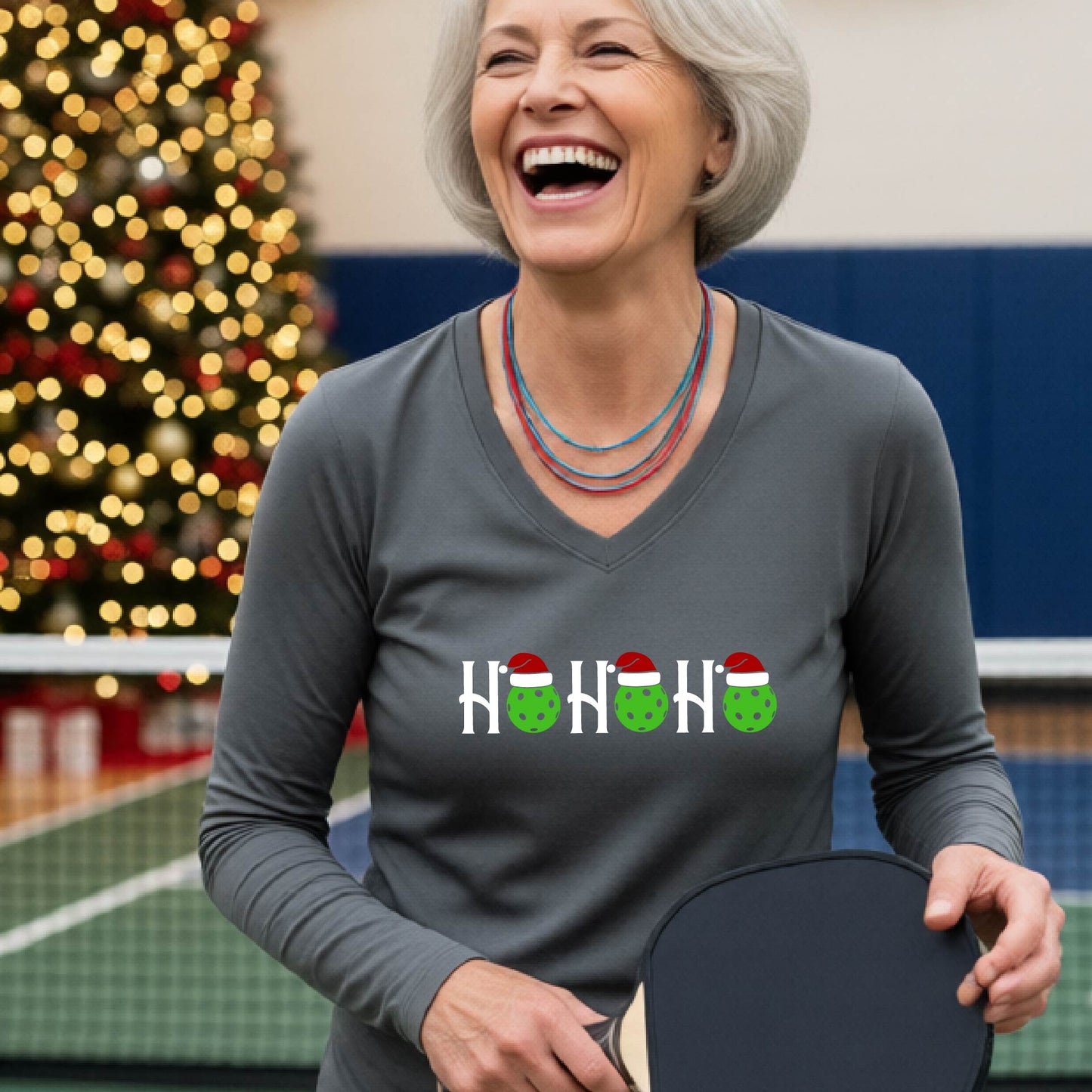 A women's gray long-sleeve v-neck shirt featuring a pickleball graphic with text "HO HO HO"  with the O replaced by green pickleballs wearing a santa hats and  Christmas themed background.