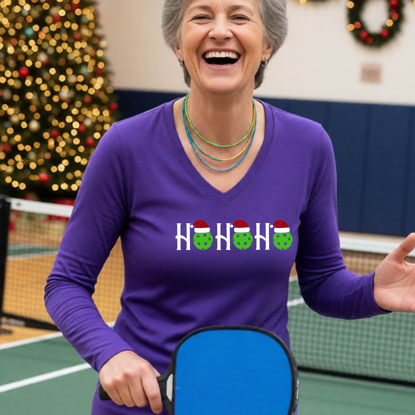 A women's purple long-sleeve v-neck shirt featuring a pickleball graphic with text "HO HO HO"  with the O replaced by green pickleballs wearing a santa hats and  Christmas themed background.