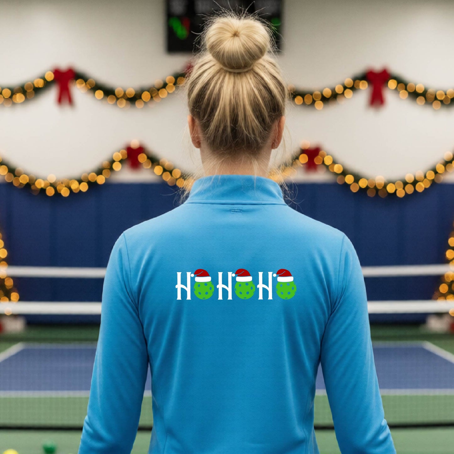 A woman viewed from behind on an indoor pickleball court, wearing a light blue long-sleeve shirt with a graphic on the back that reads 'HOHOHO' in white with green pickleballs and Santa hats.