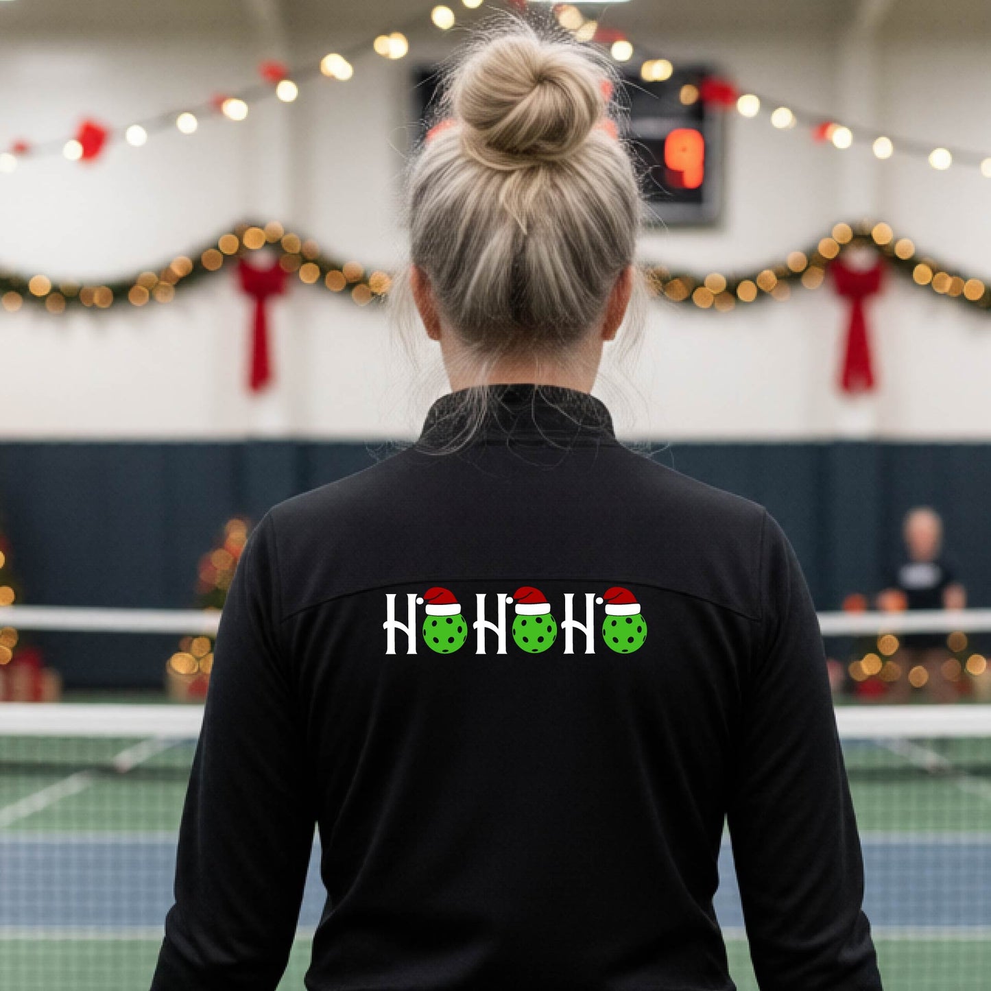 A woman viewed from behind on an indoor pickleball court, wearing a black long-sleeve shirt with a graphic on the back that reads 'HOHOHO' in white with green pickleballs and Santa hats.