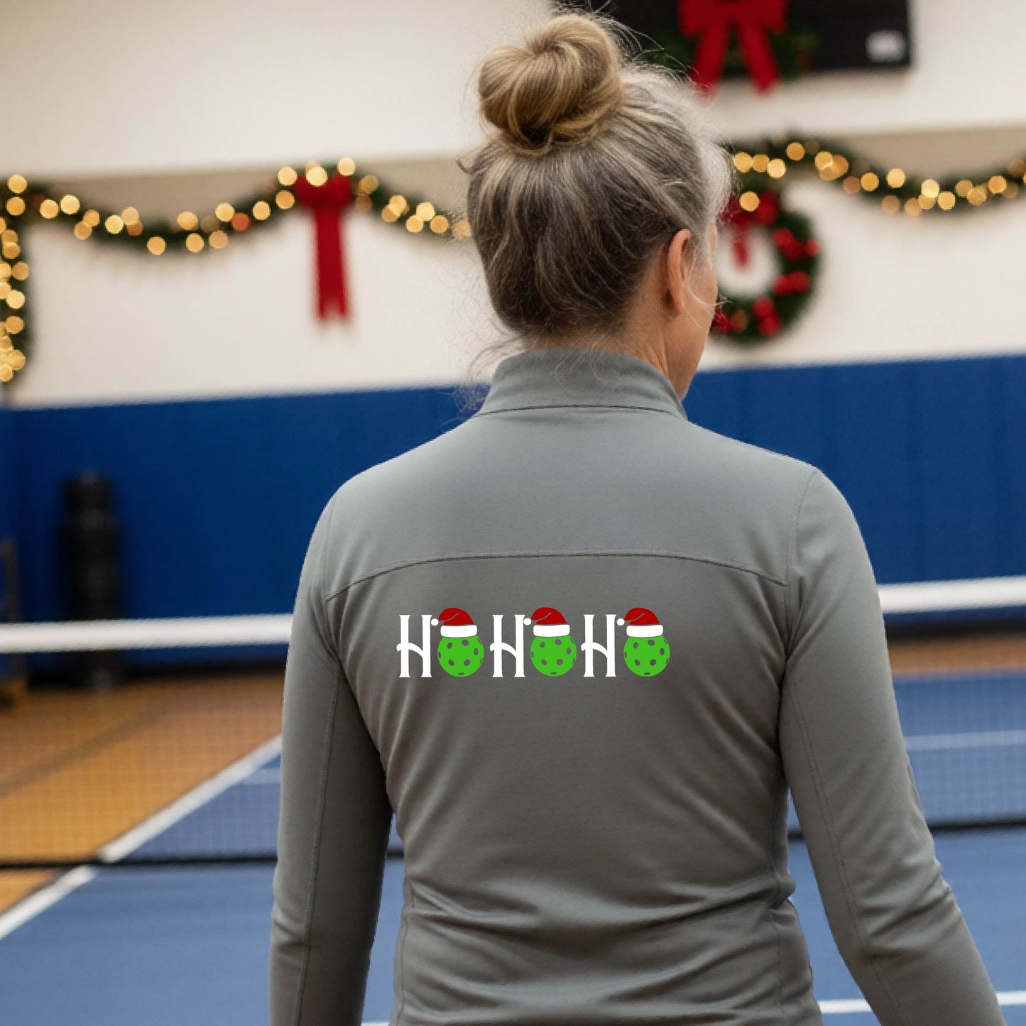 A woman viewed from behind on an indoor pickleball court, wearing a gray long-sleeve shirt with a graphic on the back that reads 'HOHOHO' in white with green pickleballs and Santa hats.