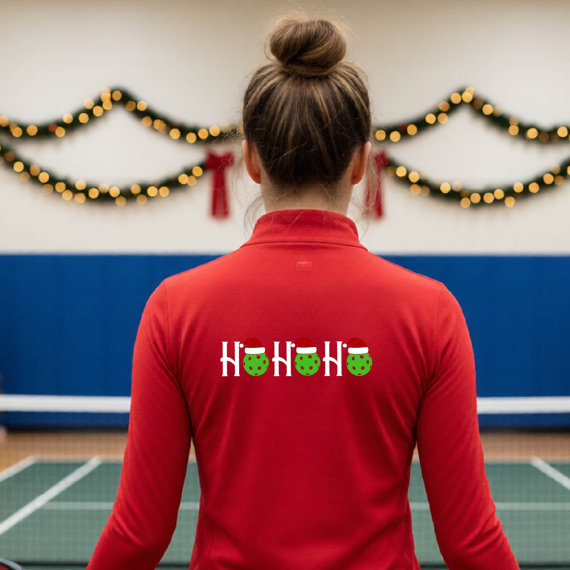 A woman viewed from behind on an indoor pickleball court, wearing a red long-sleeve shirt with a graphic on the back that reads 'HOHOHO' in white with green pickleballs and Santa hats.