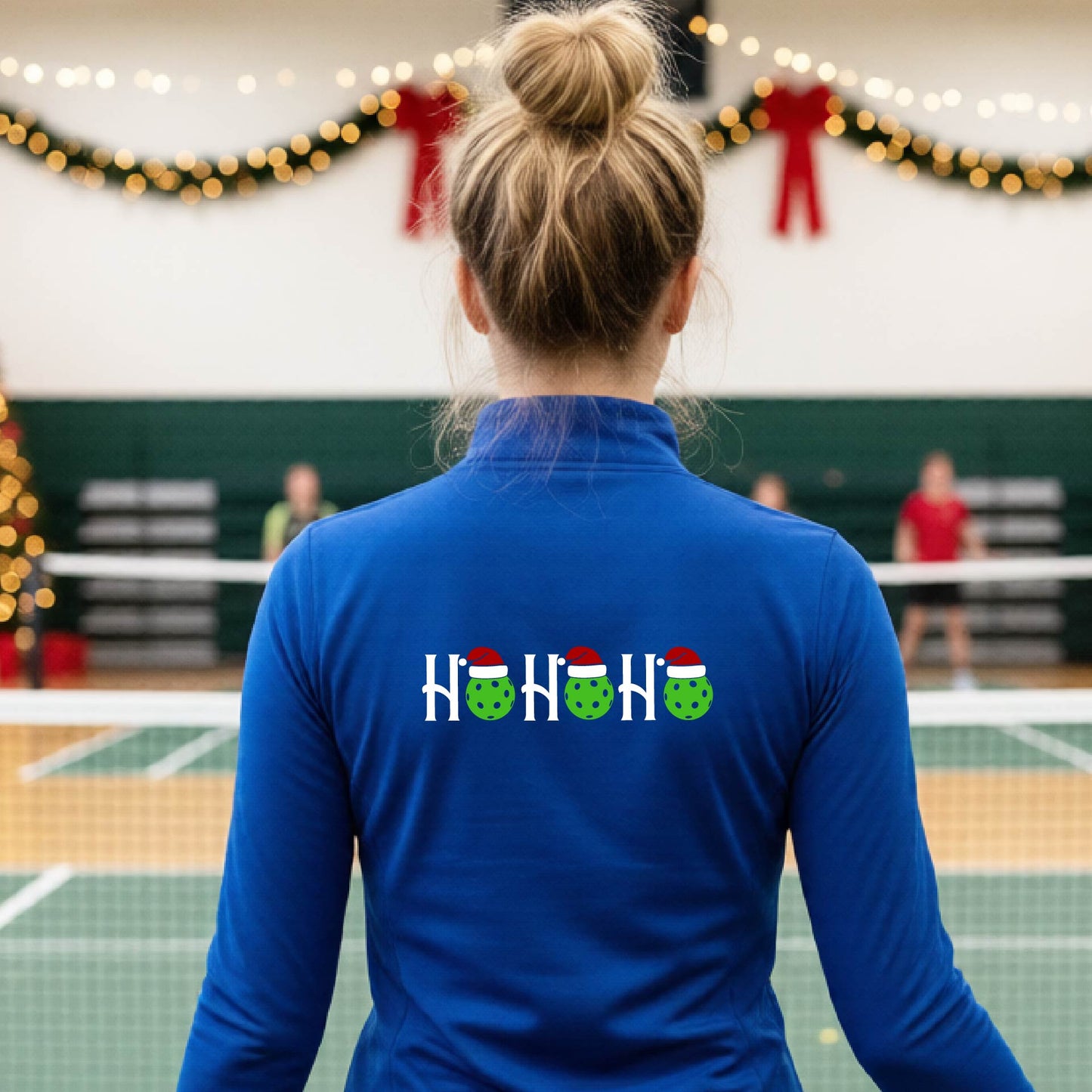 A woman viewed from behind on an indoor pickleball court, wearing a royal blue  long-sleeve shirt with a graphic on the back that reads 'HOHOHO' in white with green pickleballs and Santa hat.