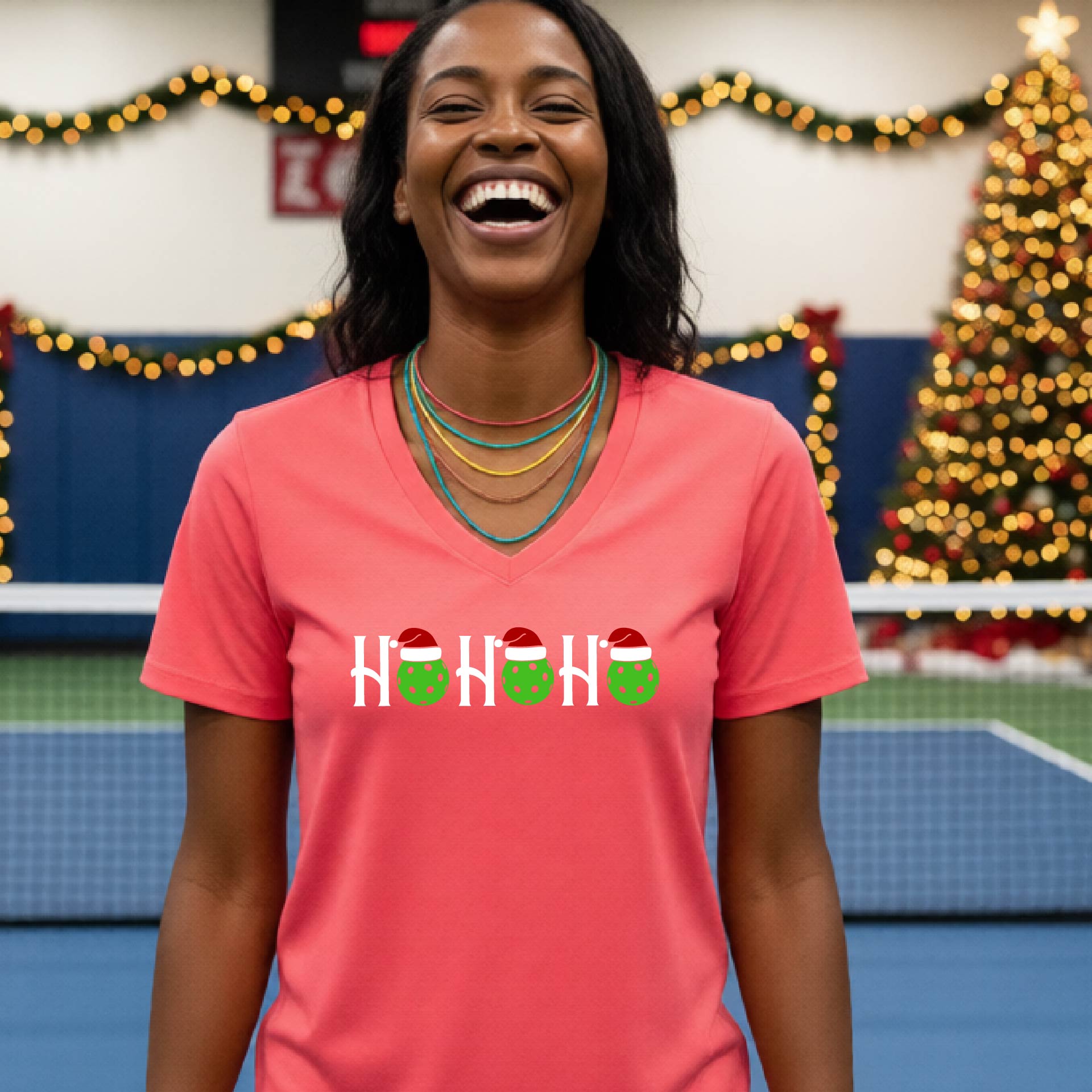 A woman smiling broadly, wearing a bright coral pink v-neck t-shirt with a graphic that reads 'HO HO HO' in white with green and red Santa hats on pickleballs. The background shows a blurred indoor tennis court with a Christmas tree and holiday lights.