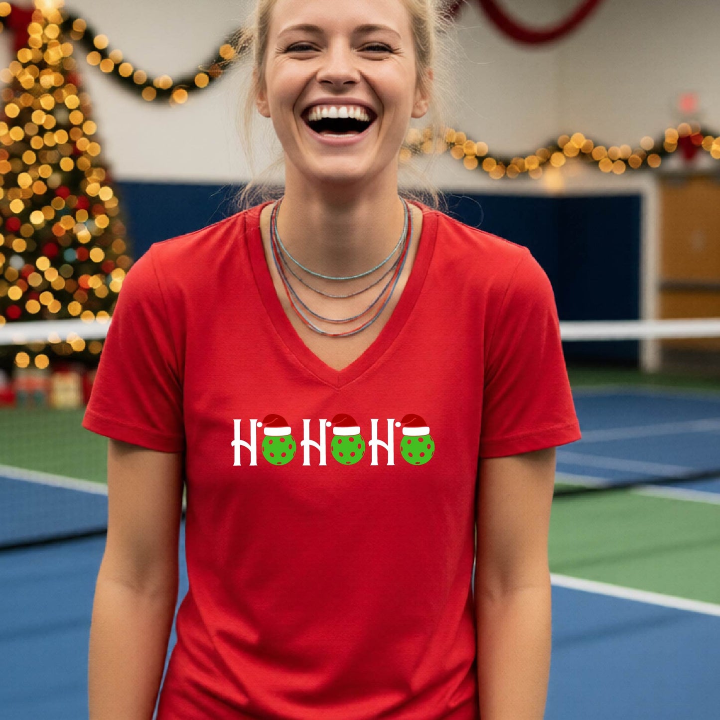 A woman smiling broadly, wearing a bright red v-neck t-shirt with a graphic that reads 'HO HO HO' in white with green and red Santa hats on pickleballs. The background shows a blurred indoor tennis court with a Christmas tree and holiday decorations.