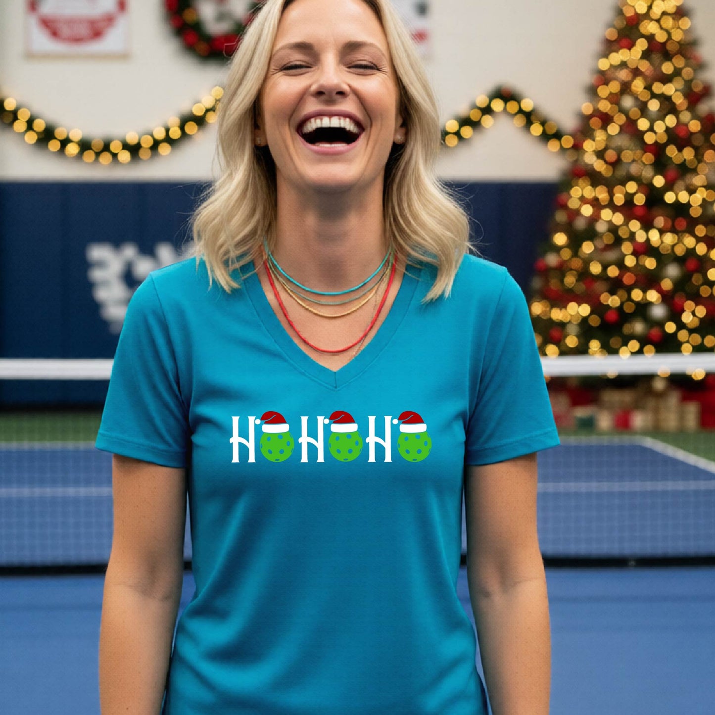 A woman smiling broadly, wearing a bright teal blue v-neck t-shirt with a graphic that reads 'HO HO HO' in white with green and red Santa hats on pickleballs. The background shows a blurred indoor tennis court with a Christmas tree and holiday lights.