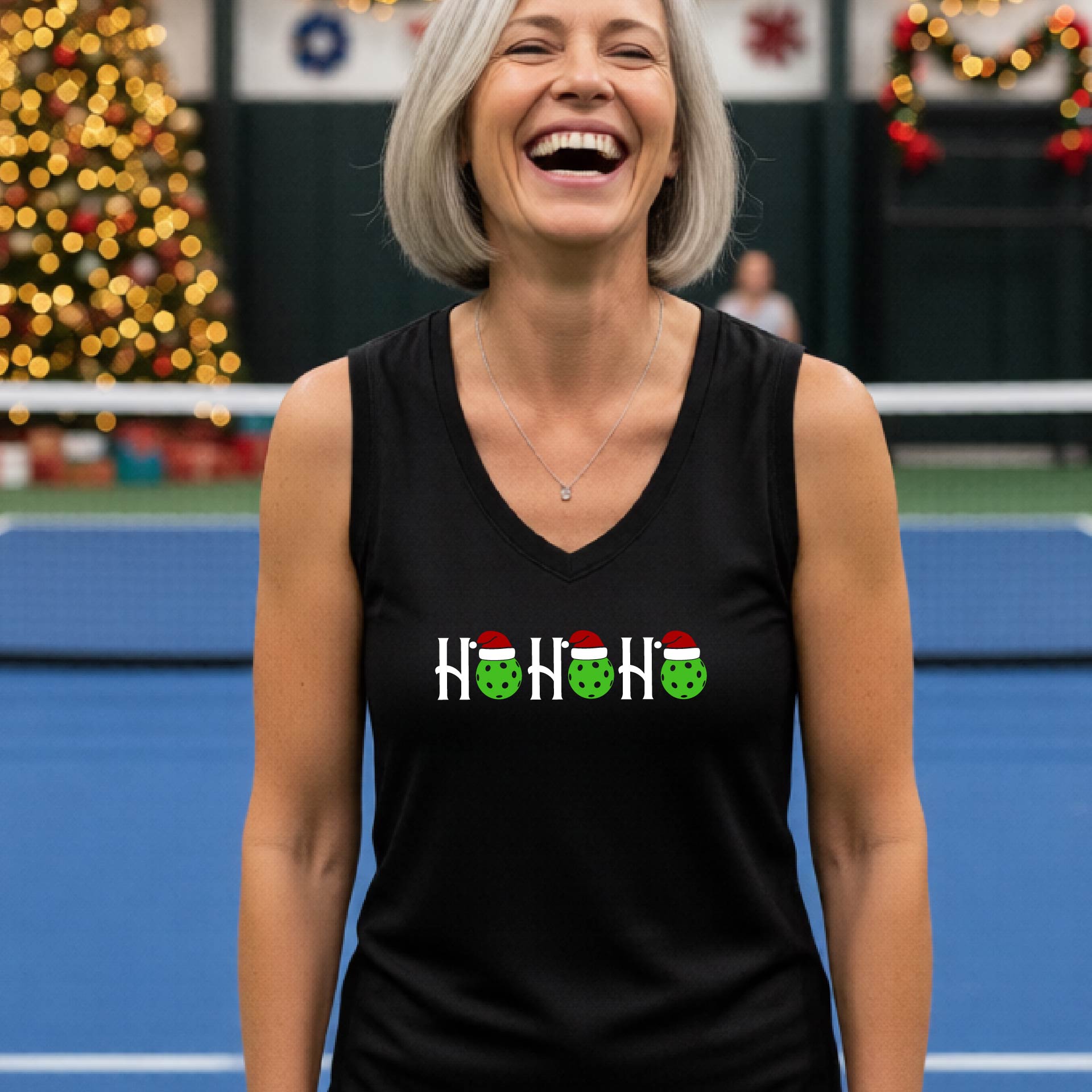 A woman laughing on a pickleball court wearing a black sleeveless tank top with a 'HO HO HO' holiday design featuring green pickleball ornaments and Santa hats. The background shows festive Christmas decorations and blurred lights.