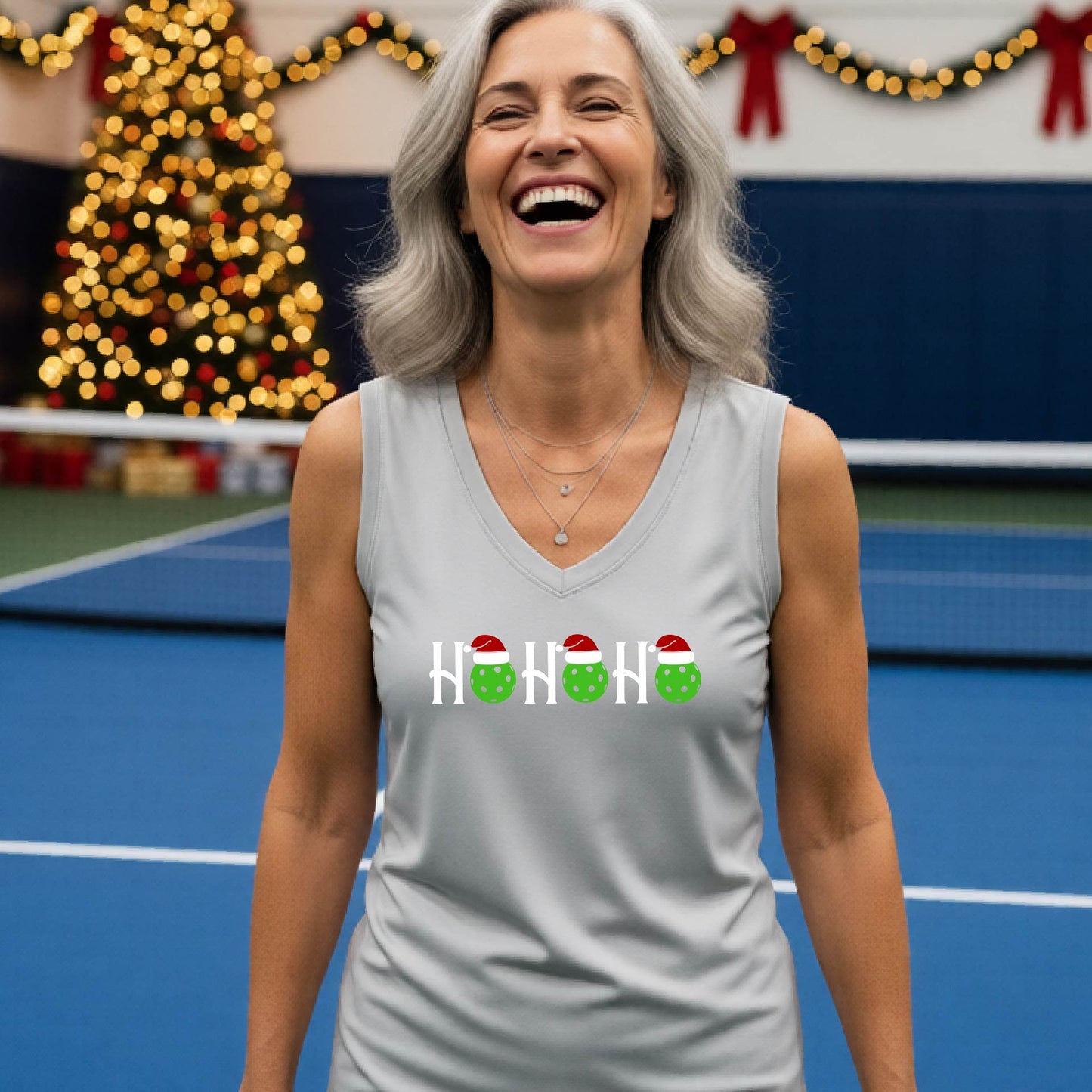 A woman laughing on a pickleball court wearing a silver gray sleeveless tank top with a 'HO HO HO' holiday design featuring green pickleball ornaments and Santa hats. The background shows festive Christmas decorations and blurred lights.