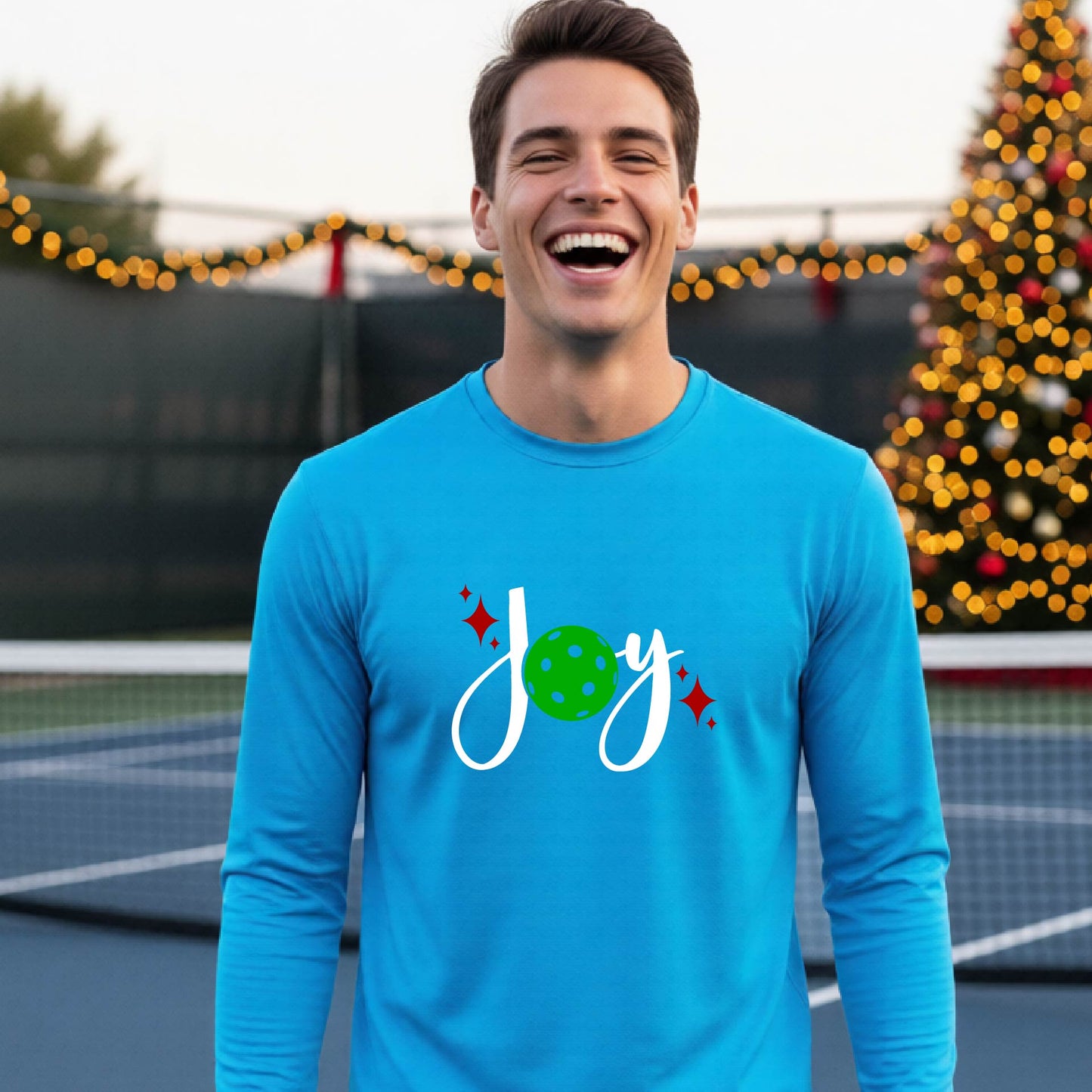 A man laughing on a pickleball court wearing a bright blue long-sleeve shirt with a 'JOY' holiday graphic with the O replaced by a green pickleball wearing a Santa hat. A decorated Christmas tree and warm string lights are blurred in the background