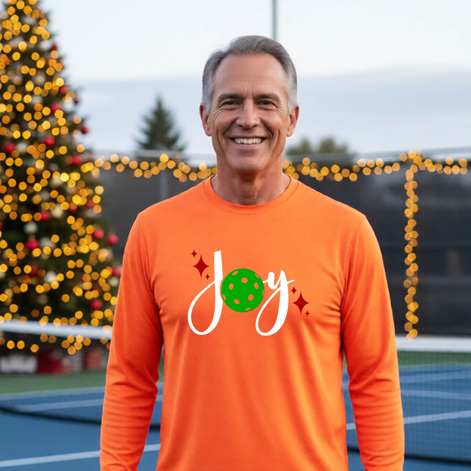 A man laughing on a pickleball court wearing a bright orange long-sleeve shirt with a 'JOY' holiday graphic with the O replaced by a green pickleball wearing a Santa hat. A decorated Christmas tree and warm string lights are blurred in the background