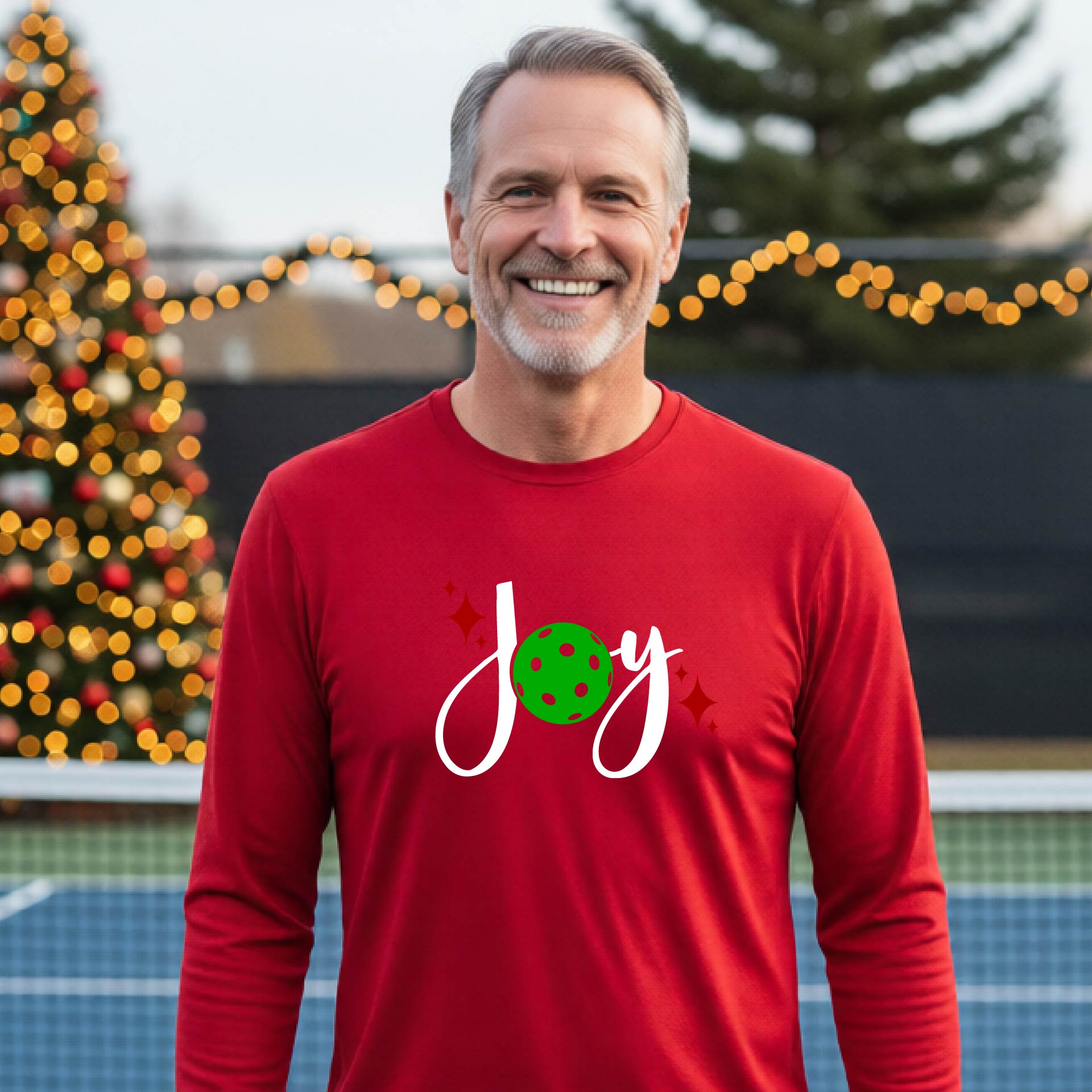 A man laughing on a pickleball court wearing a red long-sleeve shirt with a 'JOY' holiday graphic with the O replaced by a green pickleball wearing a Santa hat. A decorated Christmas tree and warm string lights are blurred in the background