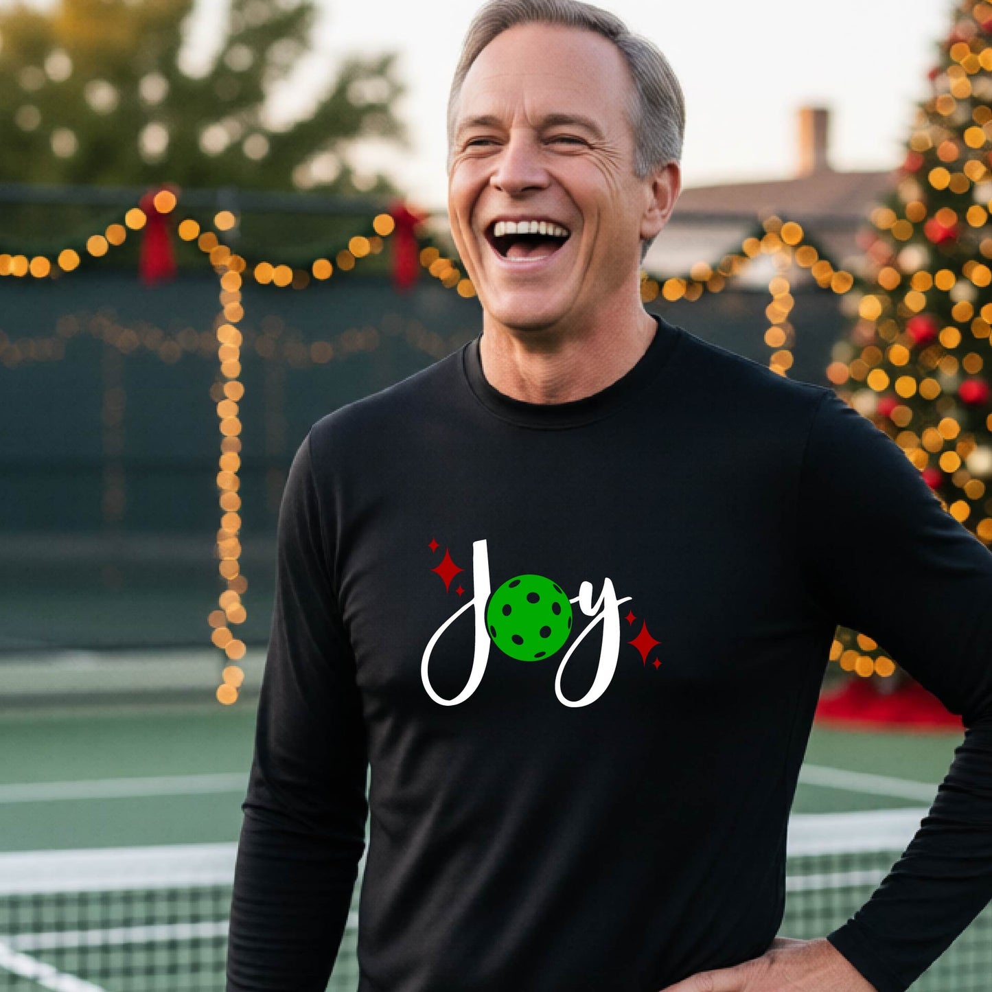 A man laughing on a pickleball court wearing a black long-sleeve shirt with a 'JOY' holiday graphic with the O replaced by a green pickleball wearing a Santa hat. A decorated Christmas tree and warm string lights are blurred in the background