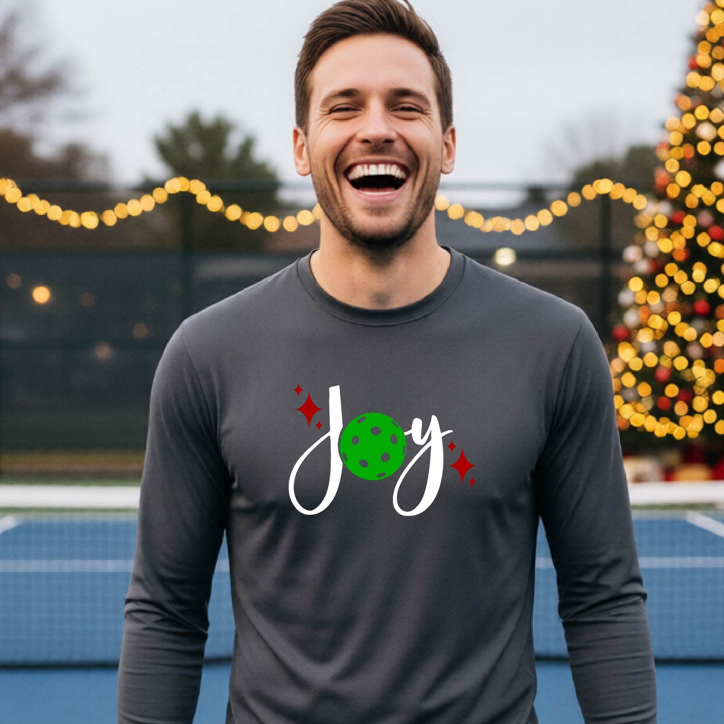 A man laughing on a pickleball court wearing a gray long-sleeve shirt with a 'JOY' holiday graphic with the O replaced by a green pickleball wearing a Santa hat. A decorated Christmas tree and warm string lights are blurred in the background