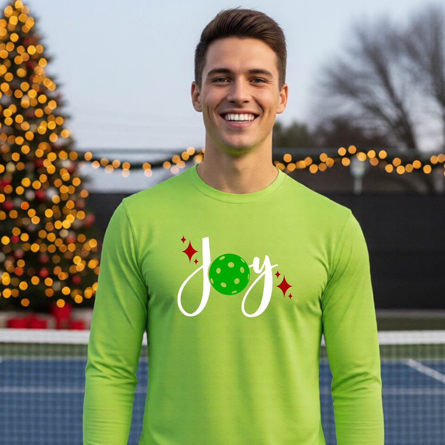 A man laughing on a pickleball court wearing a bright lime green long-sleeve shirt with a 'JOY' holiday graphic with the O replaced by a green pickleball wearing a Santa hat. A decorated Christmas tree and warm string lights are blurred in the background