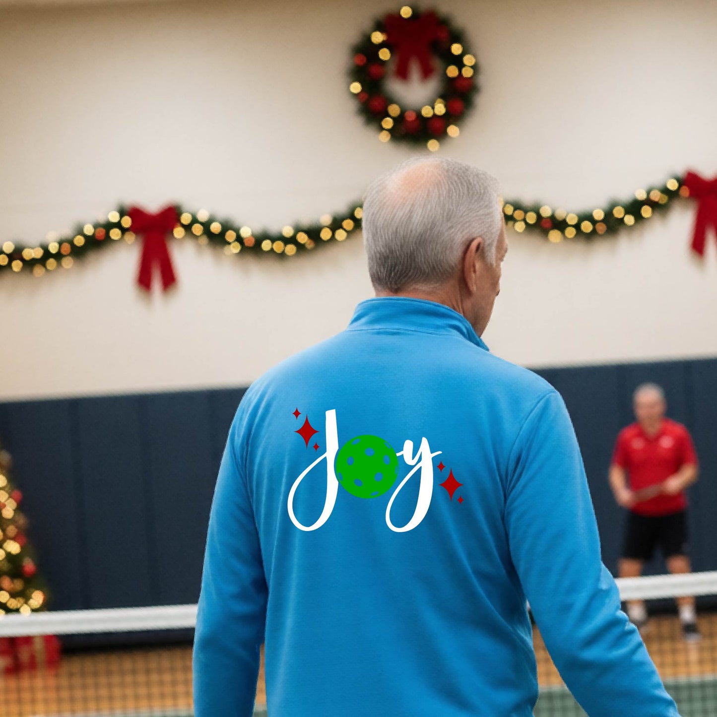 A man viewed from behind wearing a bright blue quarter-zip pullover with a festive Christmas graphic on the back that reads 'Joy' in white script with a green pickleball and red stars. The photo is taken inside a gymnasium decorated for Christmas.