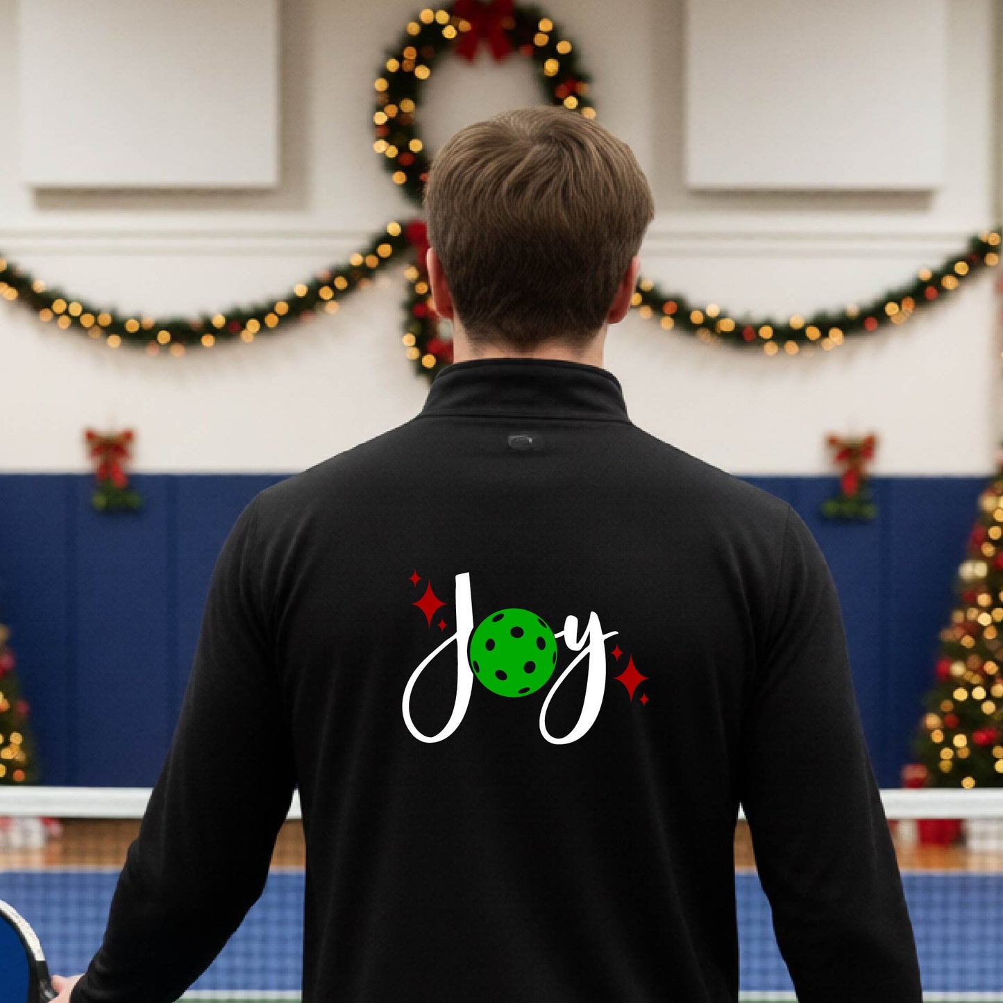 A man viewed from behind wearing a royal black quarter-zip pullover with a festive Christmas graphic on the back that reads 'Joy' in white script with a green pickleball and red stars. The photo is taken inside a gymnasium decorated for Christmas.