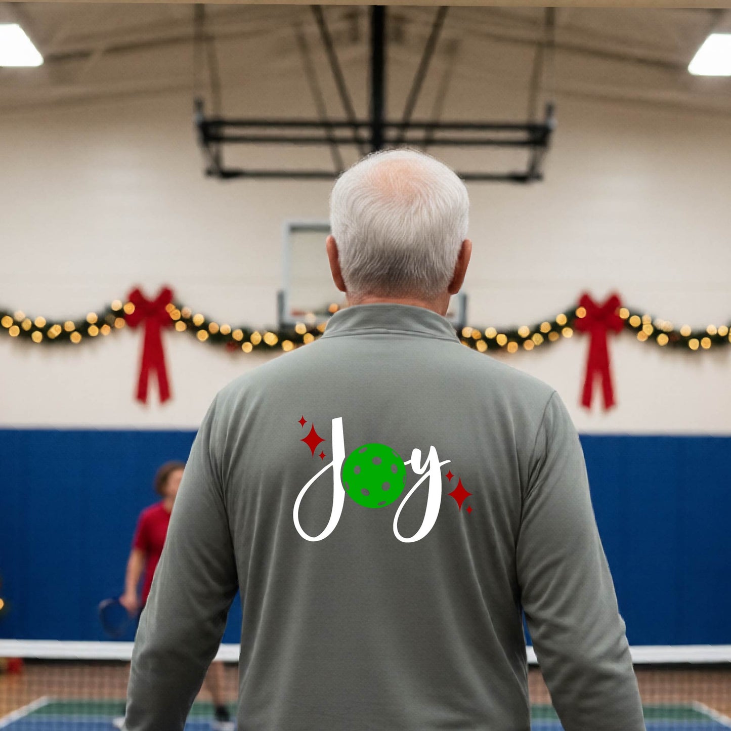 A man viewed from behind wearing a grey quarter-zip pullover with a festive Christmas graphic on the back that reads 'Joy' in white script with a green pickleball and red stars. The photo is taken inside a gymnasium decorated for Christmas.
